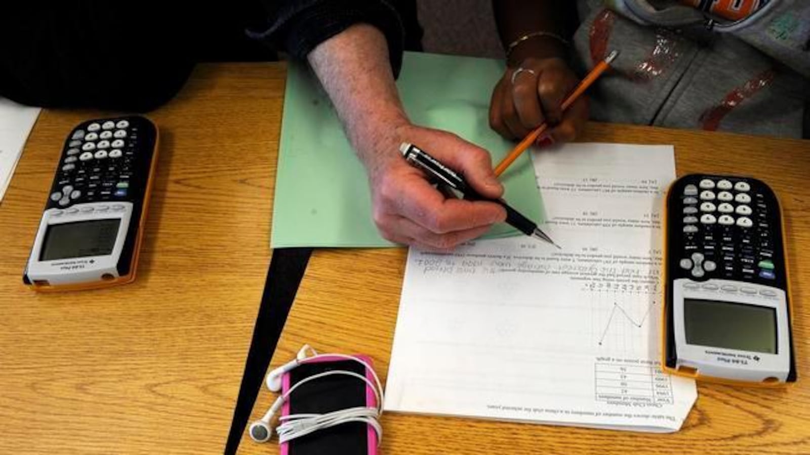 A students with a pencil writes on a white piece of paper with calculators and a green piece of paper nearby.