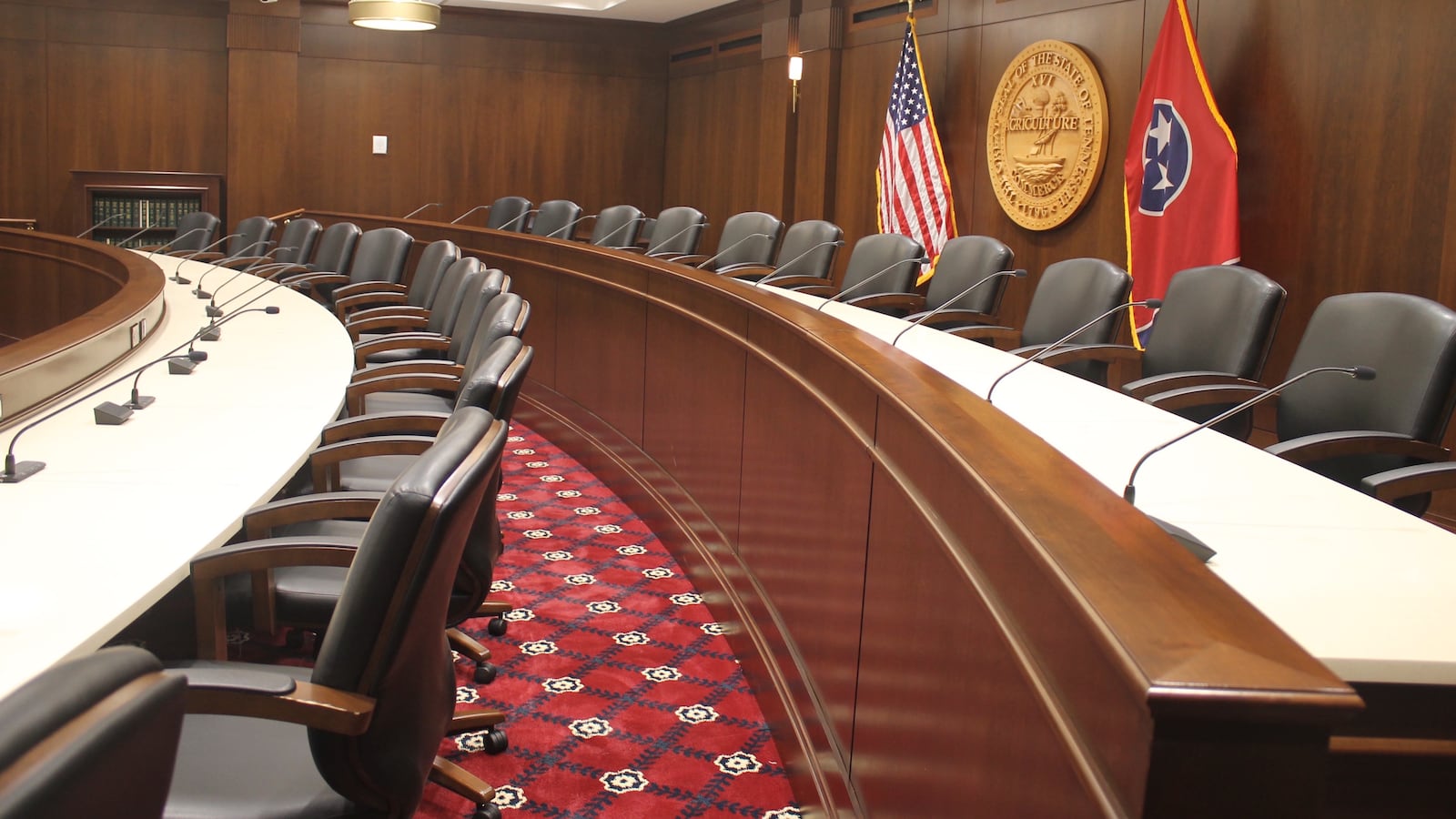 An empty legislative chambers with two rows of empty chairs with an American and Tennessee flag in the background with the state seal.
