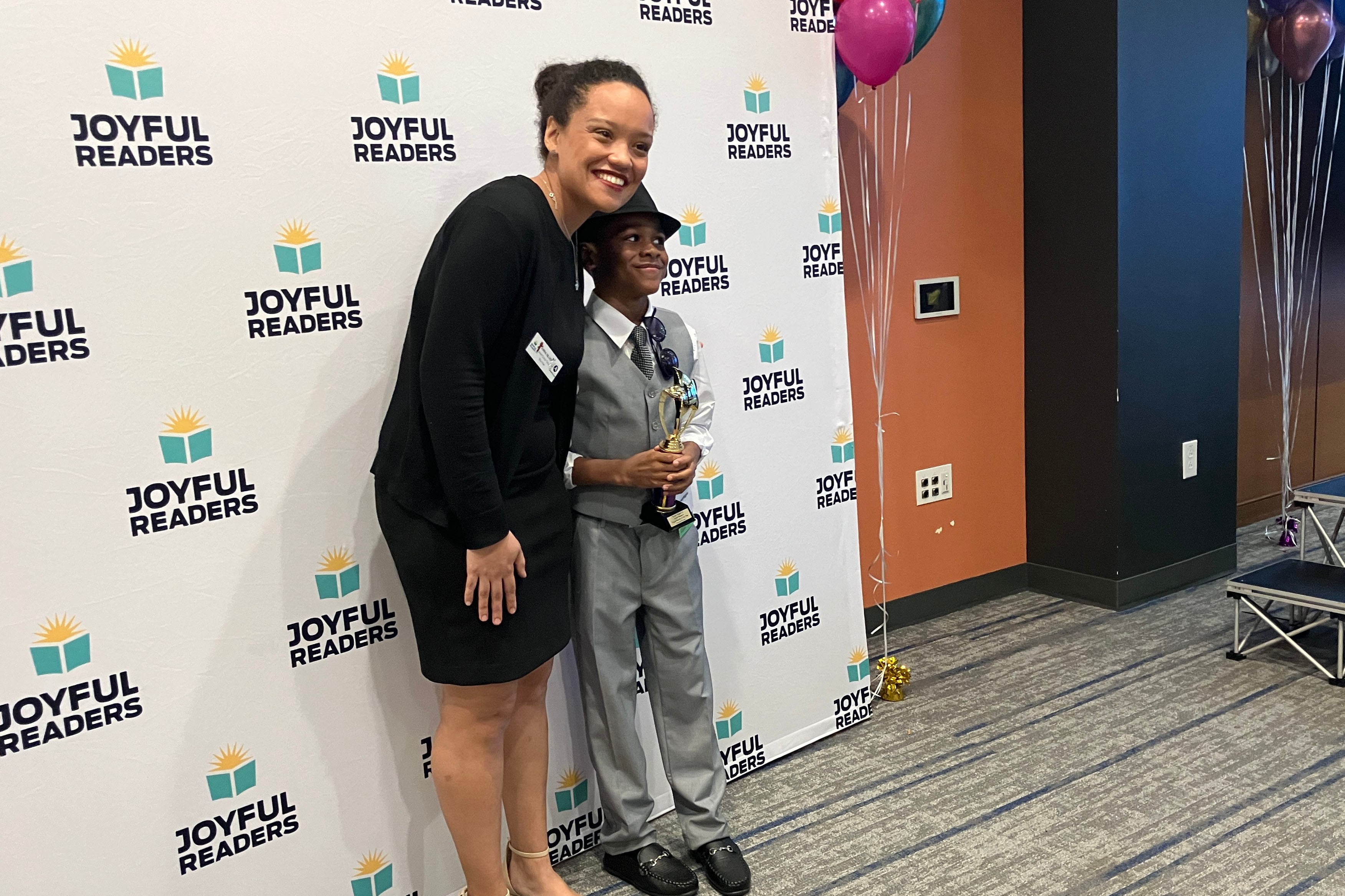An adult woman wearing a black dress poses with a young Black boy in a grey suit and wearing a fedora holds a trophy in front of a white background with a colorful logo.