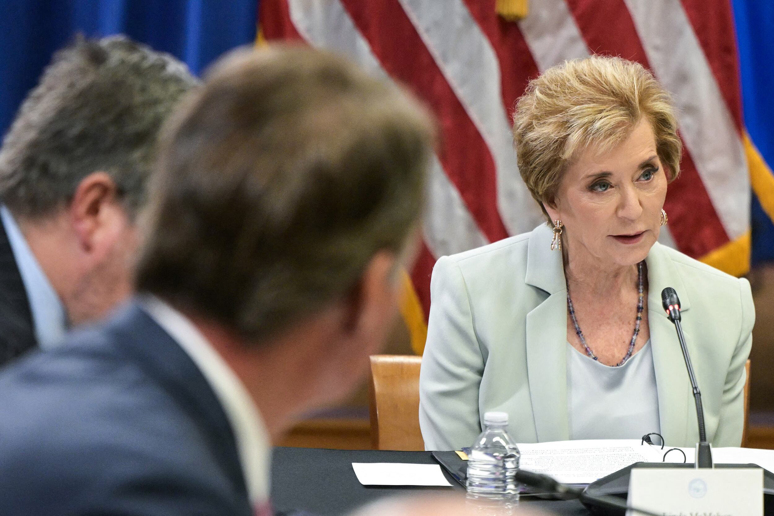 A photograph of an older white woman in a light blue suit speaks into a thin microphone while sitting down and the backs of two people's heads are in the foreground.