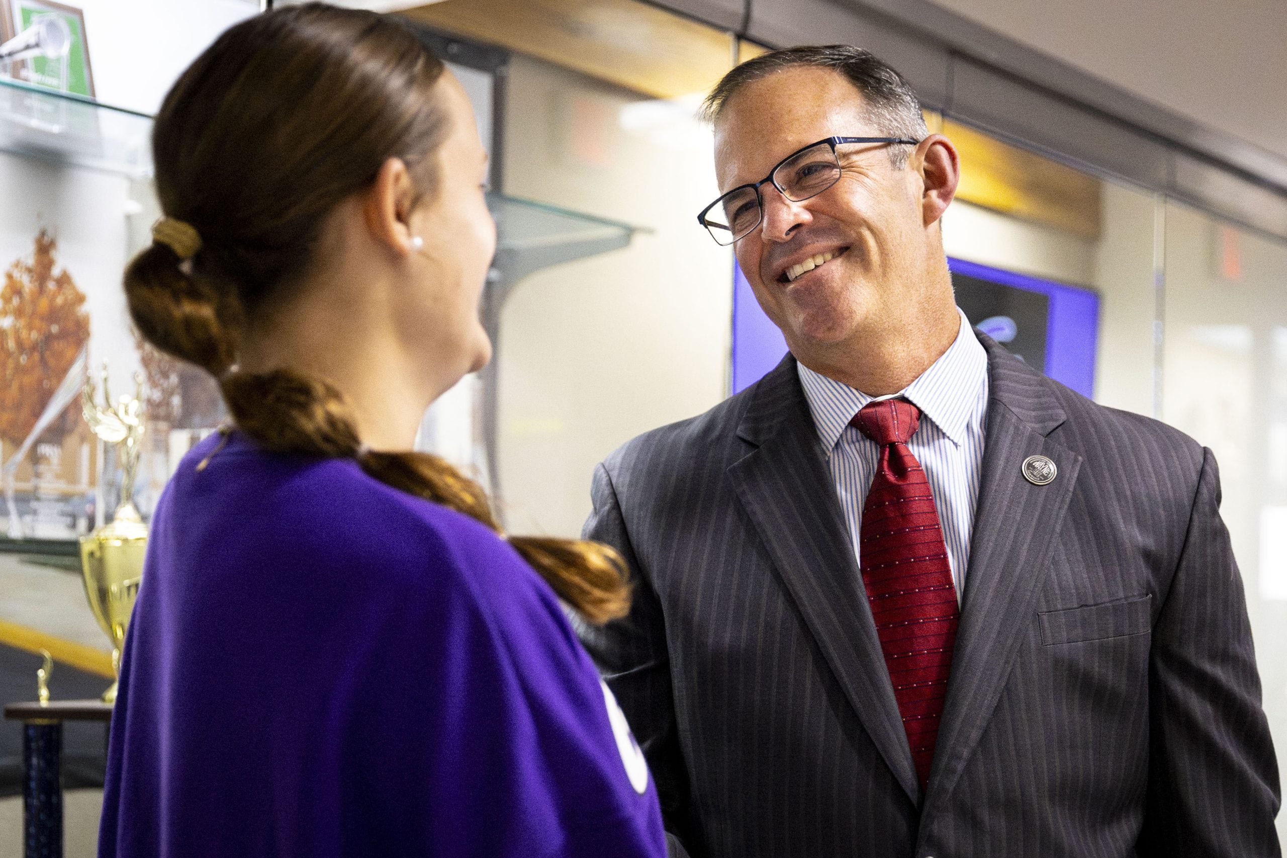 A photograph of a white man in a suit standing next to and talking with a white female in a purple sweater.