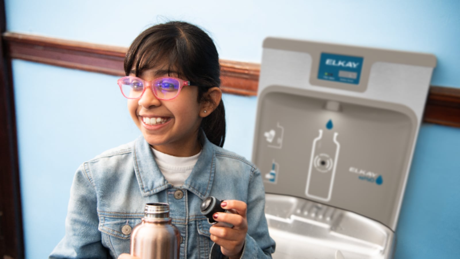 A girl smiles after filling her water bottle at a filtrated water fountain; she is in a school setting.