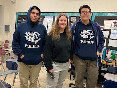 Two high school students wearing dark hoodies with their school logo pose with their high school teacher who has long light brown hair and is wearing a black sweater. The three are posing side by side in a classroom.