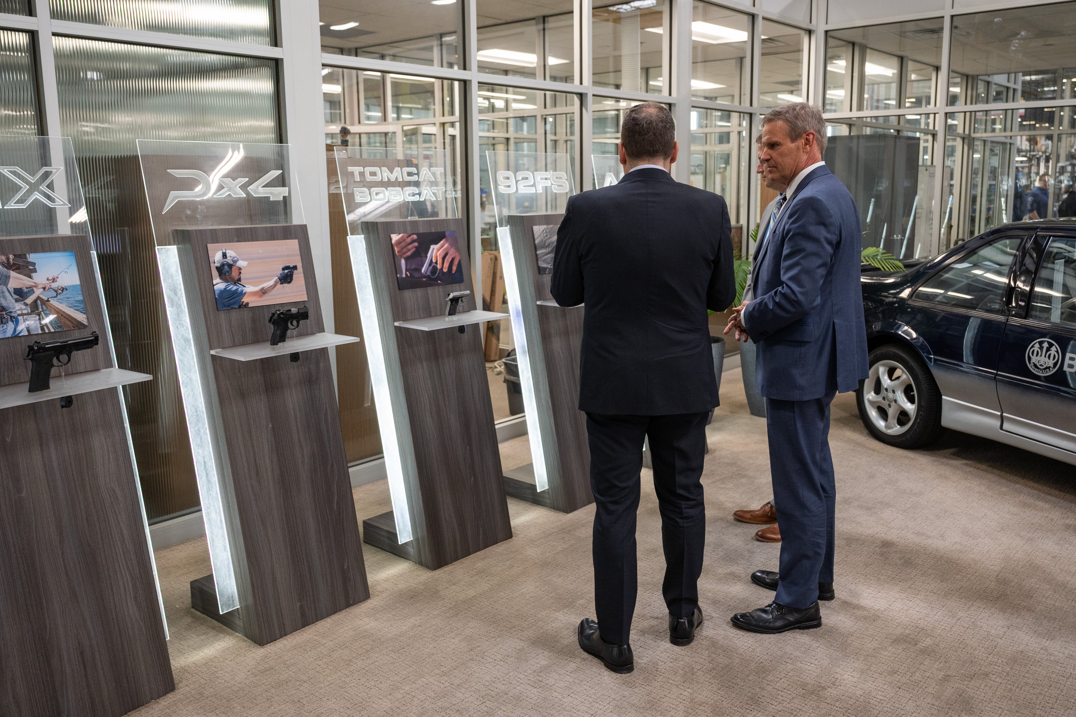 A man wearing a suit looks at a display of handguns with two other men in a factory setting.