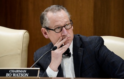 A man in a suit sits at a wooden desk with a microphone and a name plaque in front of his face.