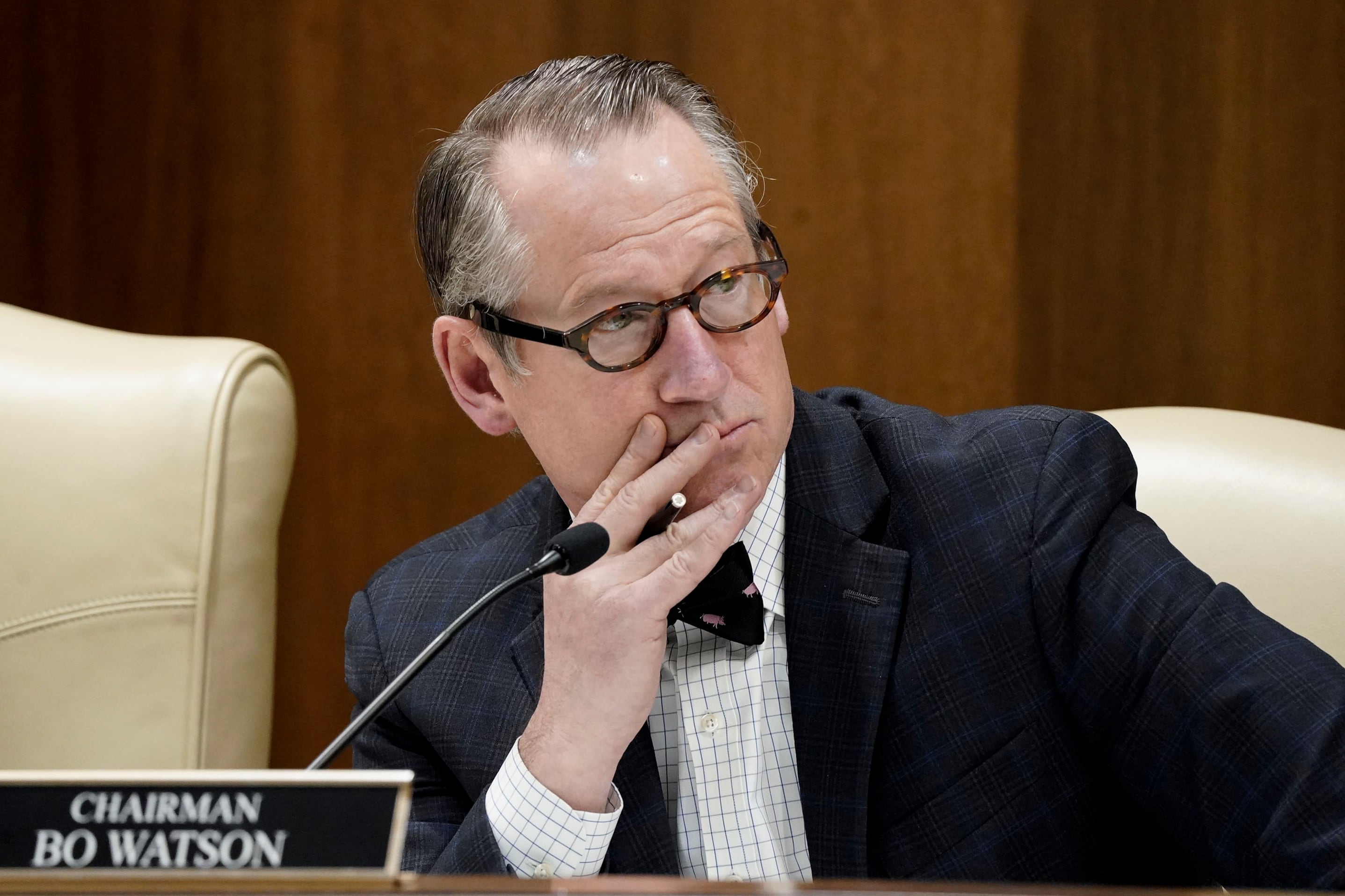 A man in a suit sits at a wooden desk with a microphone and a name plaque in front of his face.