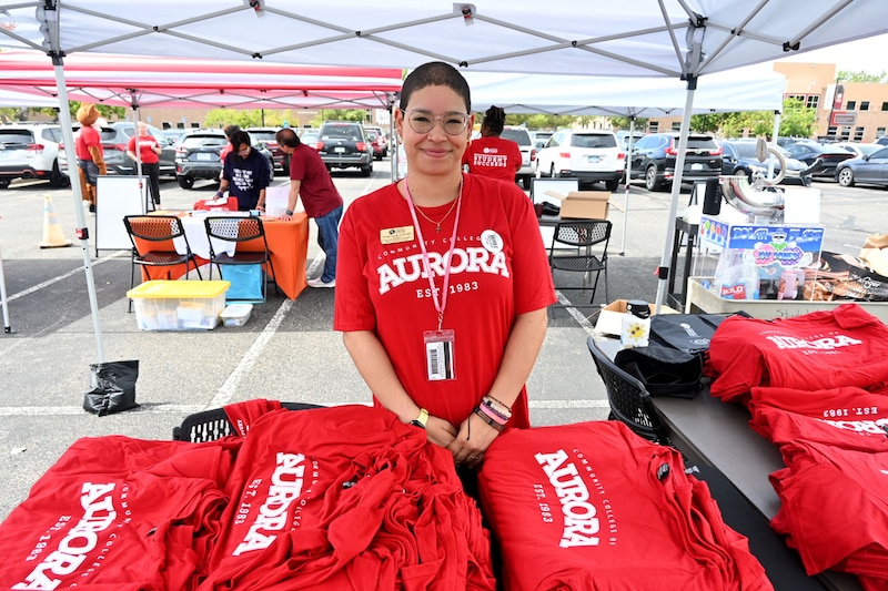 An adult poses for a portrait next to a table full of red t-shirts and stands under a temporary tent outside.