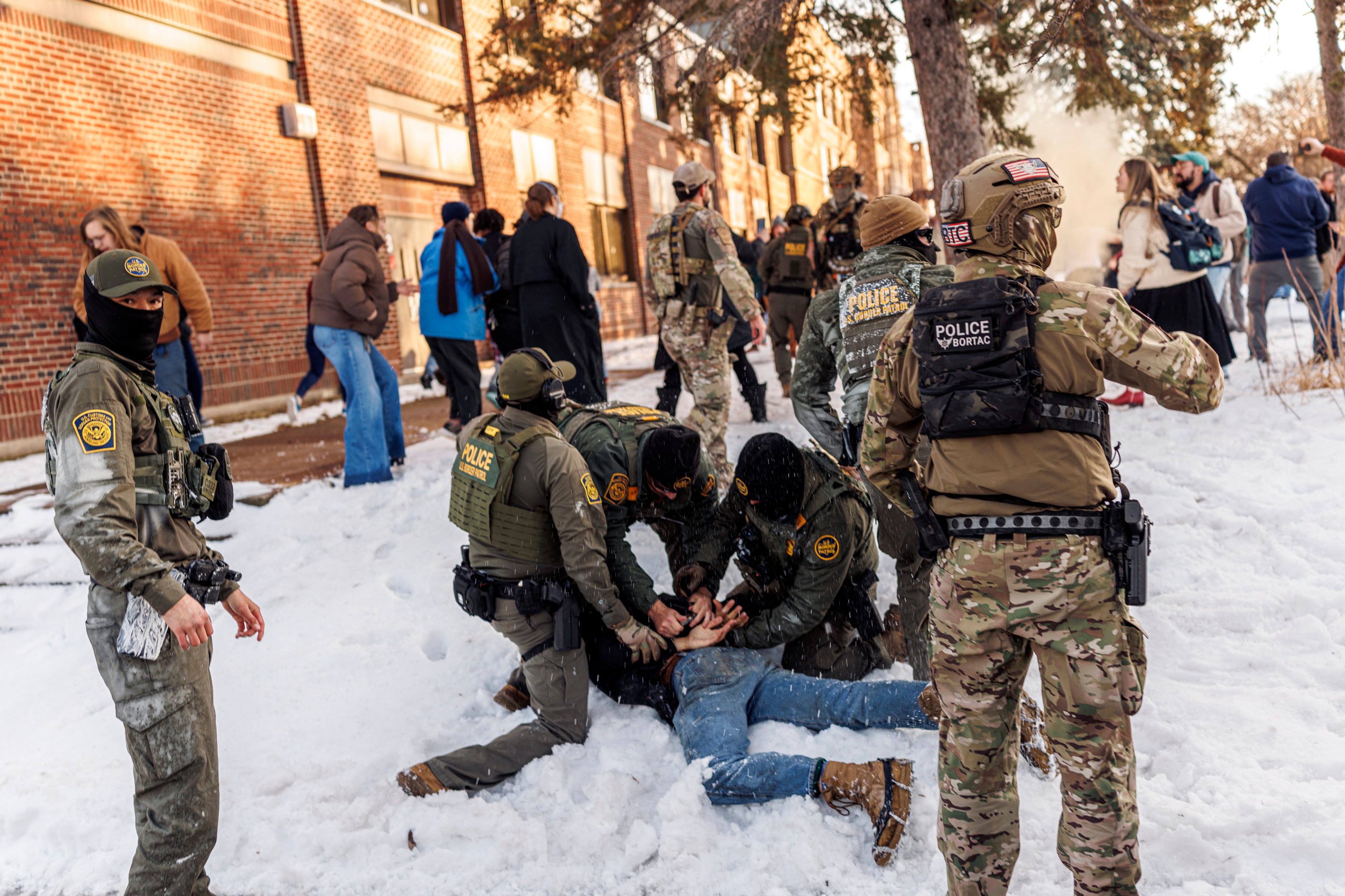 A photograph of a group of armed ICE agents tackling a person who is face down in the snow with their hands behind their backs.