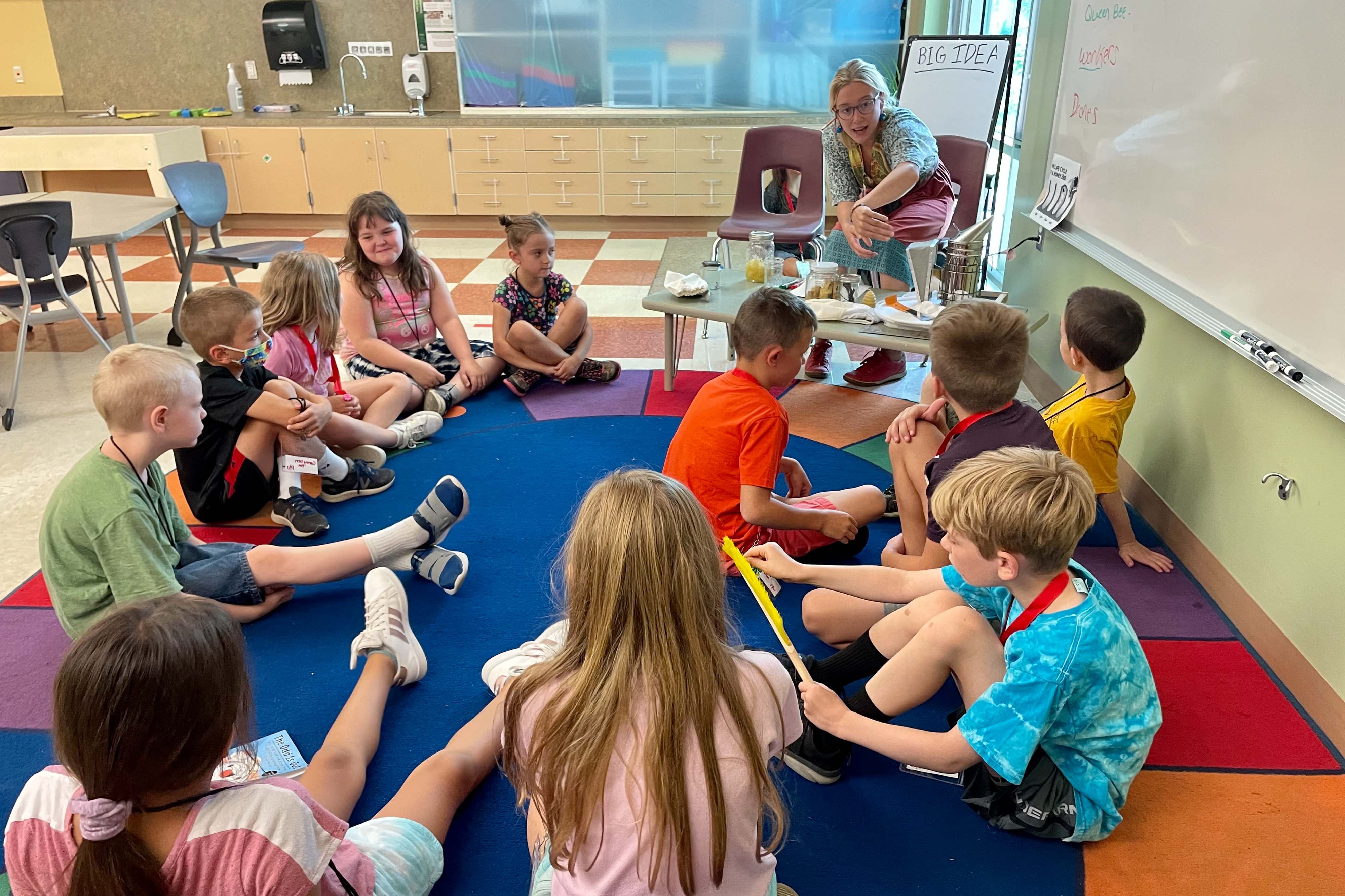 A teacher shows her students beekeeping equipment in the classroom, as the children sit on a large, multi-colored rug.