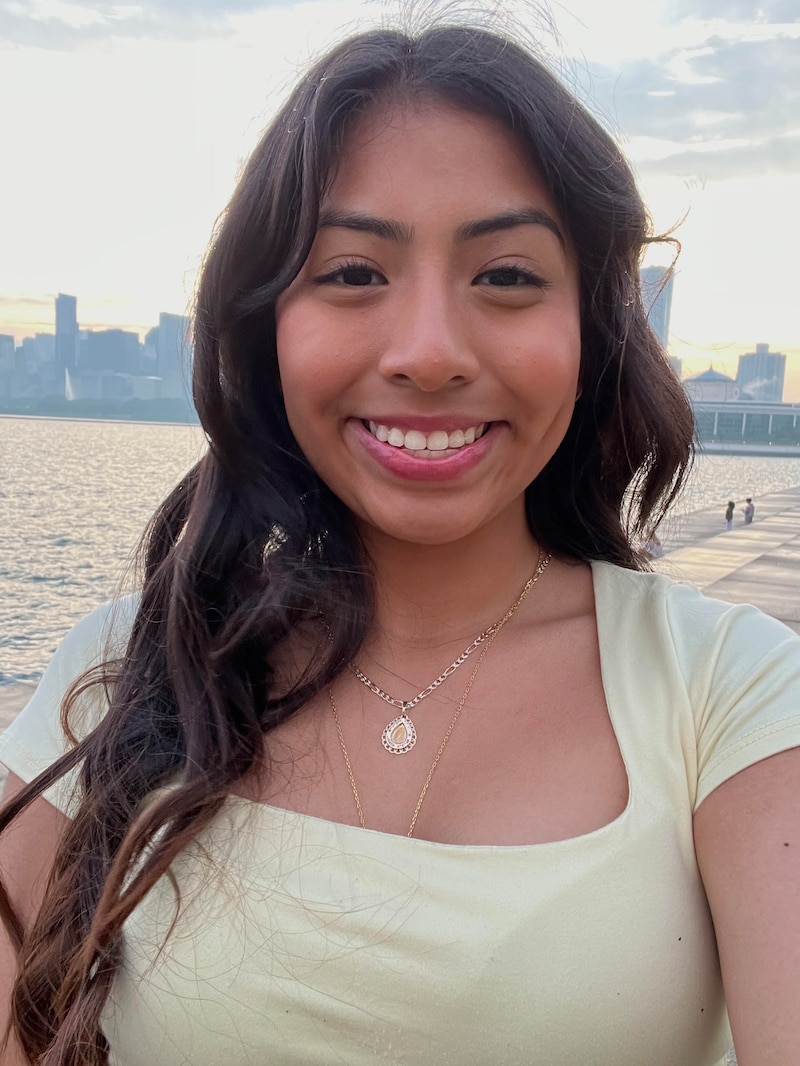 A high school senior girl with long dark hair and wearing a yellow blouse poses for a portrait in front of a lake view with a skyline in the background.