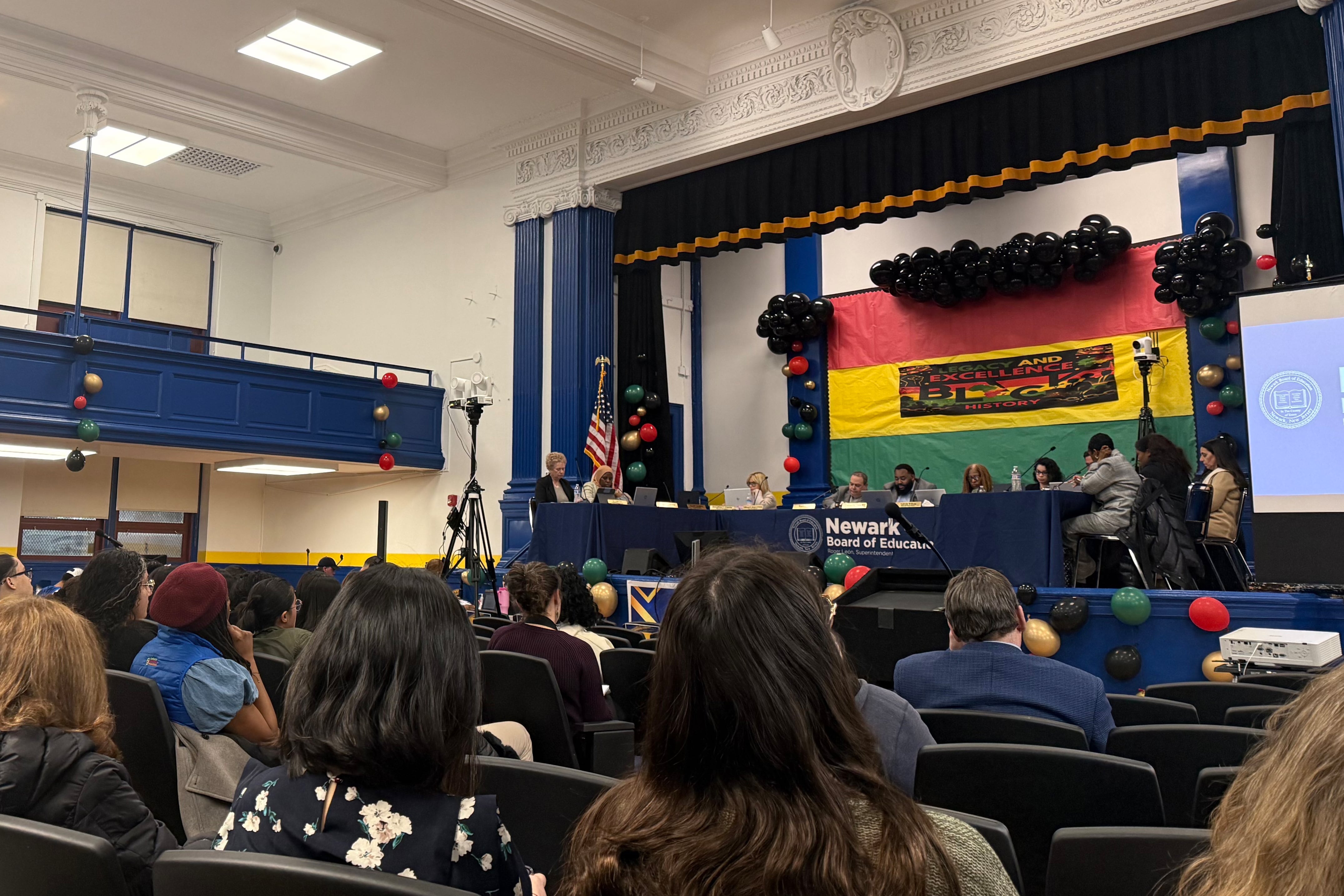 A photograph of adults sitting on stage during a school board meeting with adults sitting in the many rows of the school auditorium.
