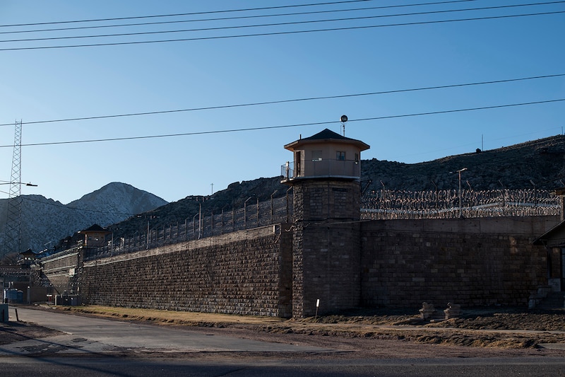 Barbed wire surrounds the top of a brick fence with a watch tower in the front and a blue sky, electrical wire and mountains in the background.