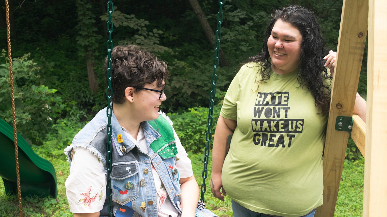 A youth in jeans and a jeans vest sits on a swing next to a person in a yellowish t-shirt that reads “Hate won’t make us great.”