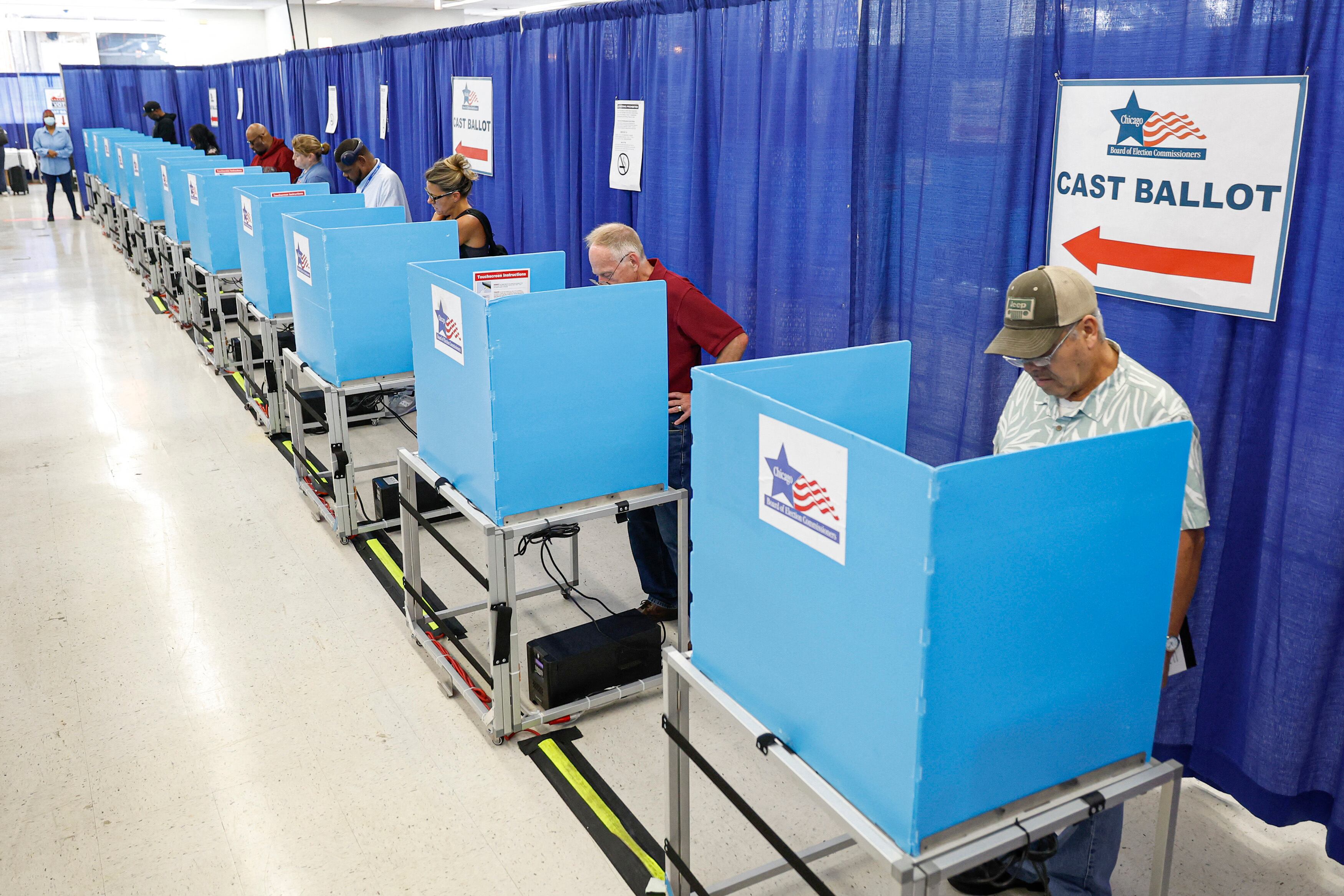A row of people stand behind small blue dividers to vote in a long room with w long blue curtain in the background.
