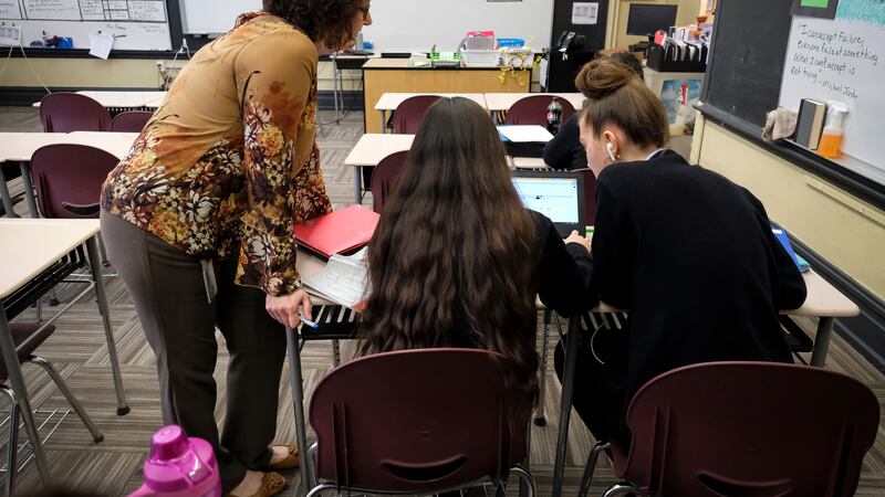 In a classroom, two students work together on a laptop while a teacher looks on.