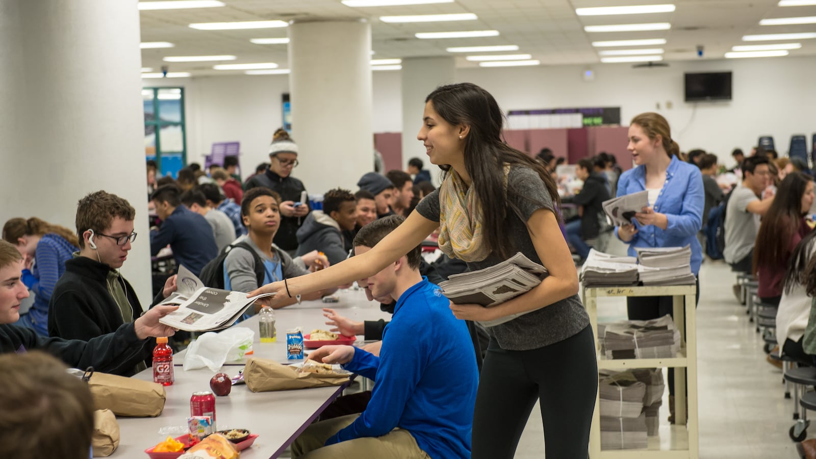 Student journalists hand out copies of a school newspaper inside a school cafeteria.