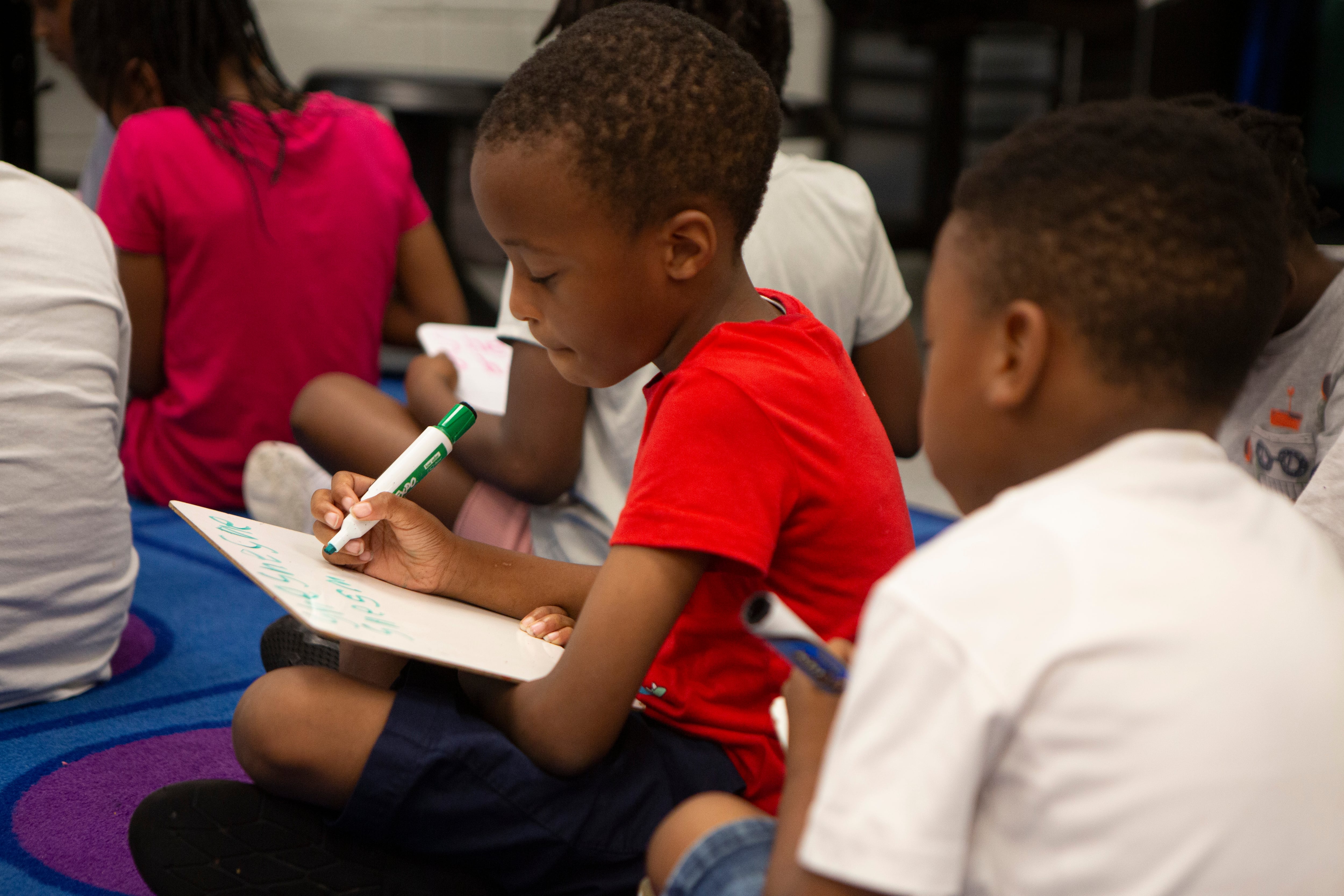 A group of young students sit on the ground in their classroom working on small dry erase boards.