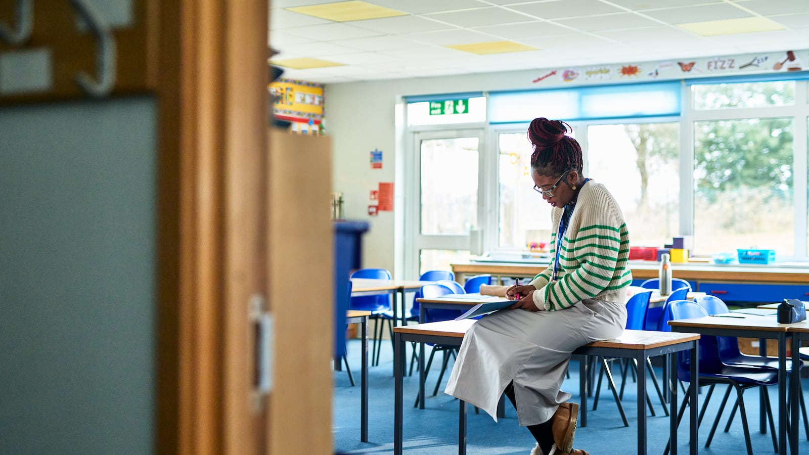 A teacher wearing a white skirt and a green and white sweater sits on a desk in an empty elementary school classroom.