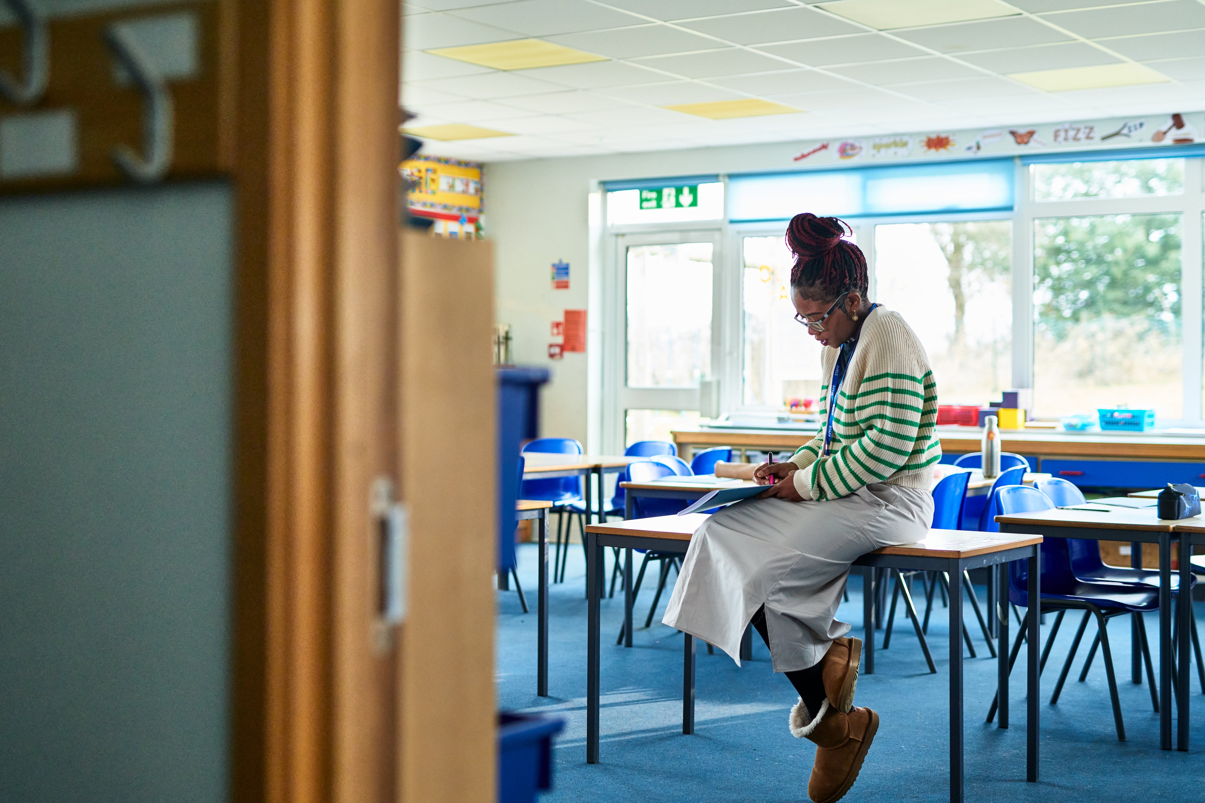 A teacher wearing a white skirt and a green and white sweater sits on a desk in an empty elementary school classroom.