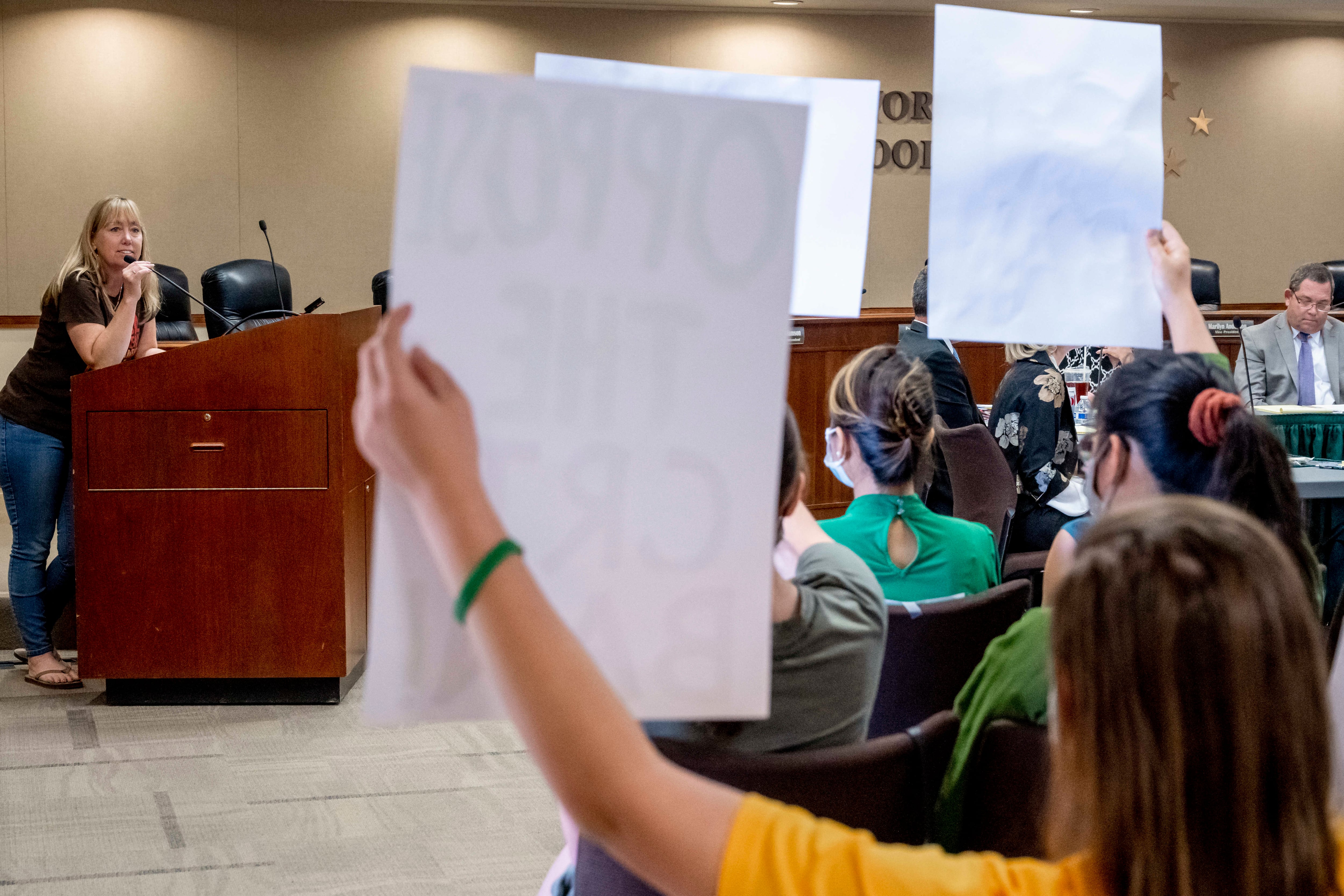 A person holds up a sign in the foreground, a person at a lectern in the background.