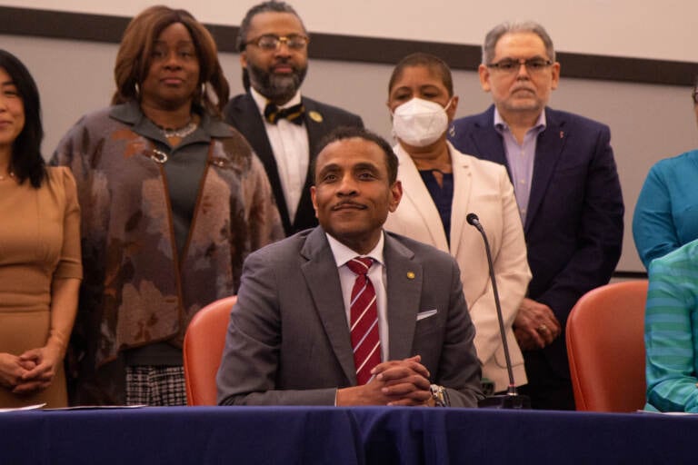 Tony Watlington sits in a gray suit and red tie at a table in front of a group of people.