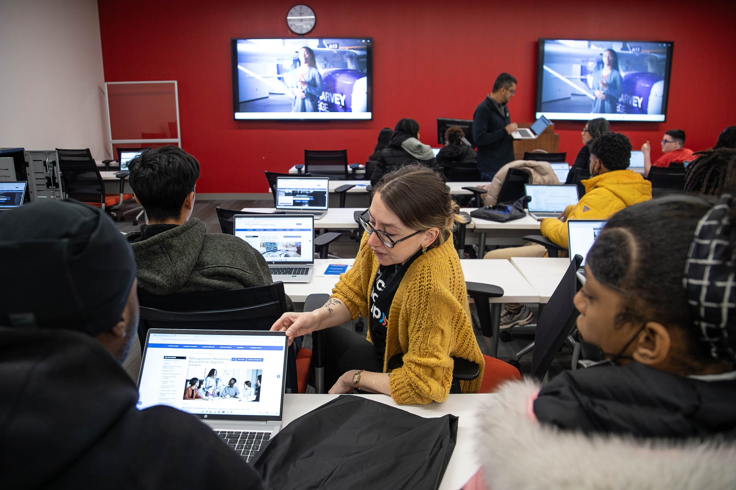 An adult and students sit at rows of tables with computers at each seat.