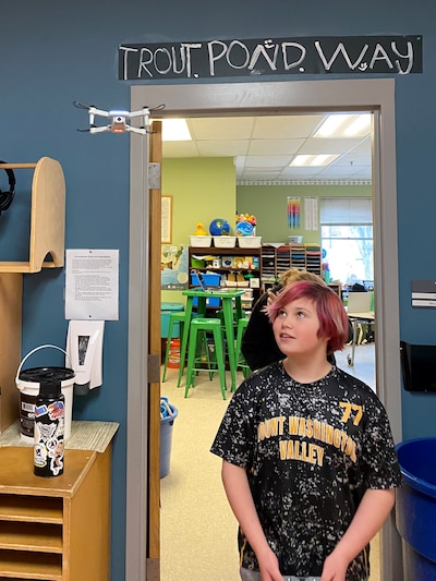 A middle school student flies a drone inside of a classroom while standing in front of a door.