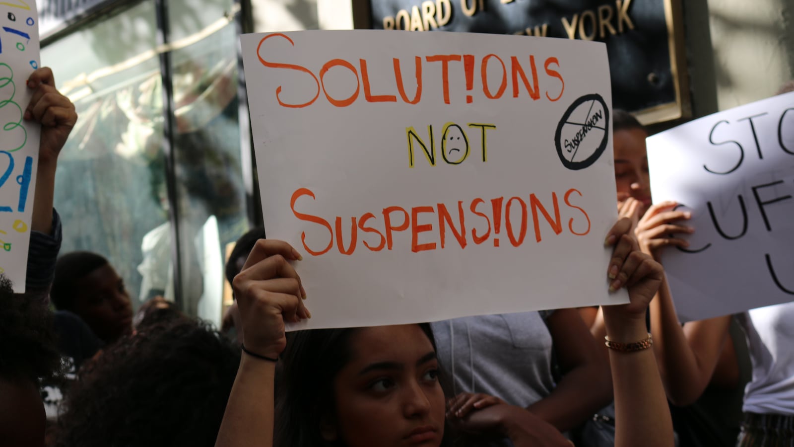 A person holds up a white sign that says Solutions Not Suspensions in orange and black letters.