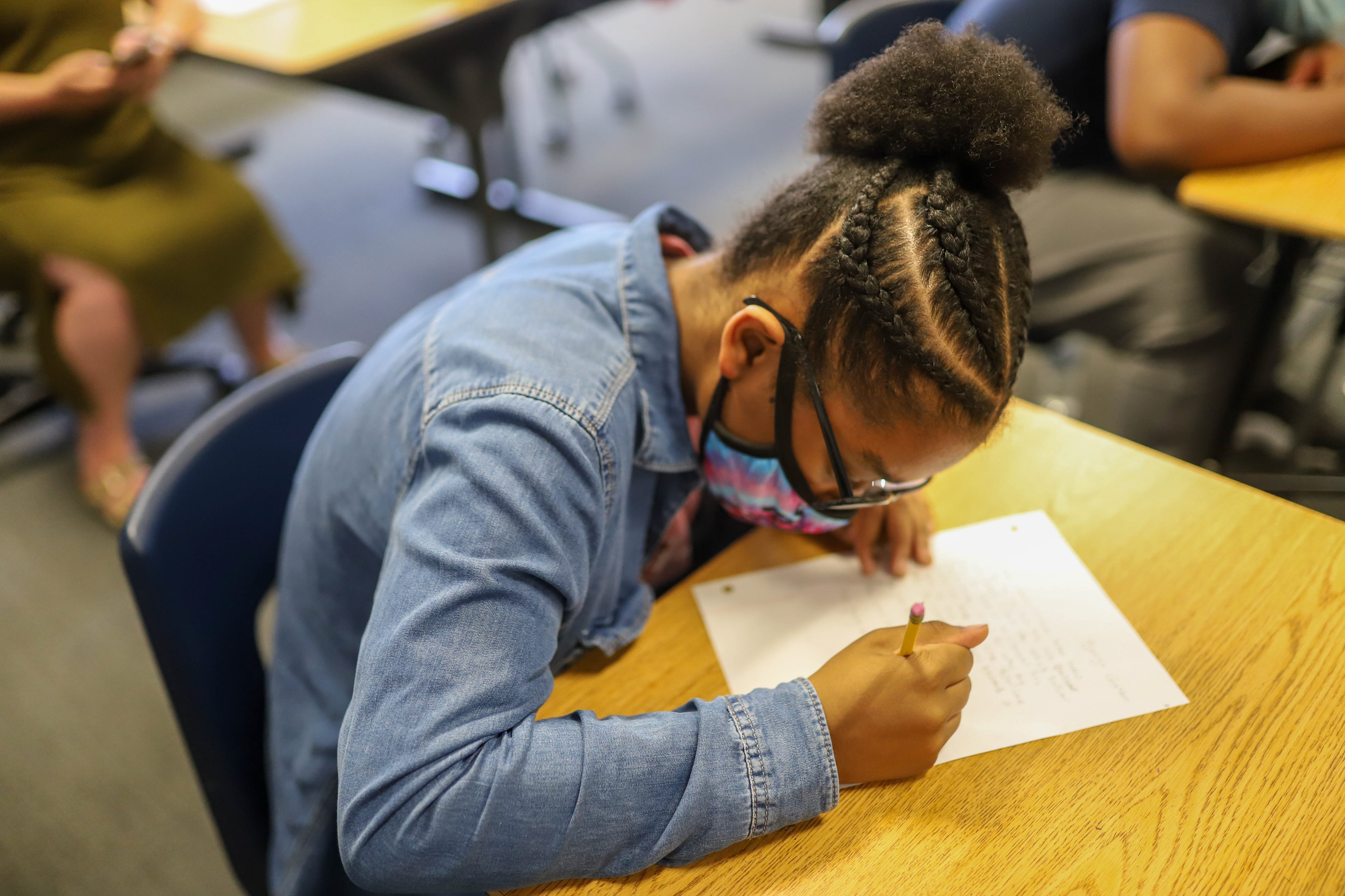 A student wearing a jean jacket and protective mask writes on a piece of notebook paper at her desk.