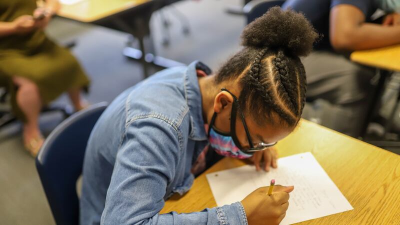 A student wearing a jean jacket and protective mask writes on a piece of notebook paper at her desk.