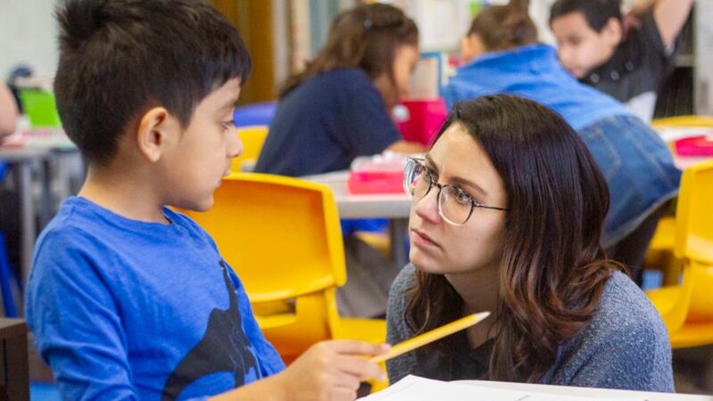Teacher Sarah Schielke, wearing glasses and blue top, crouches down to listen to a student, who is sitting at a desk and holding a yellow pencil.