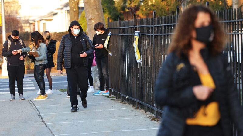Teens wearing puffy coats and masks walk by a fence in front of a school.