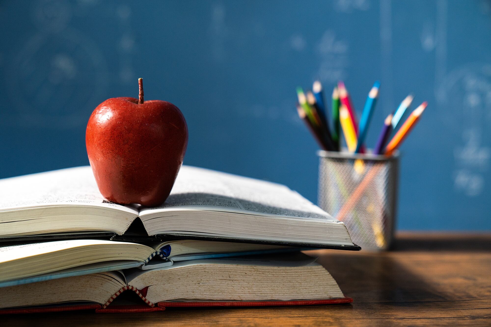 A red apple sits perfectly on top of three opened books and in the background there is a colored pencil holder in front of a chalkboard.