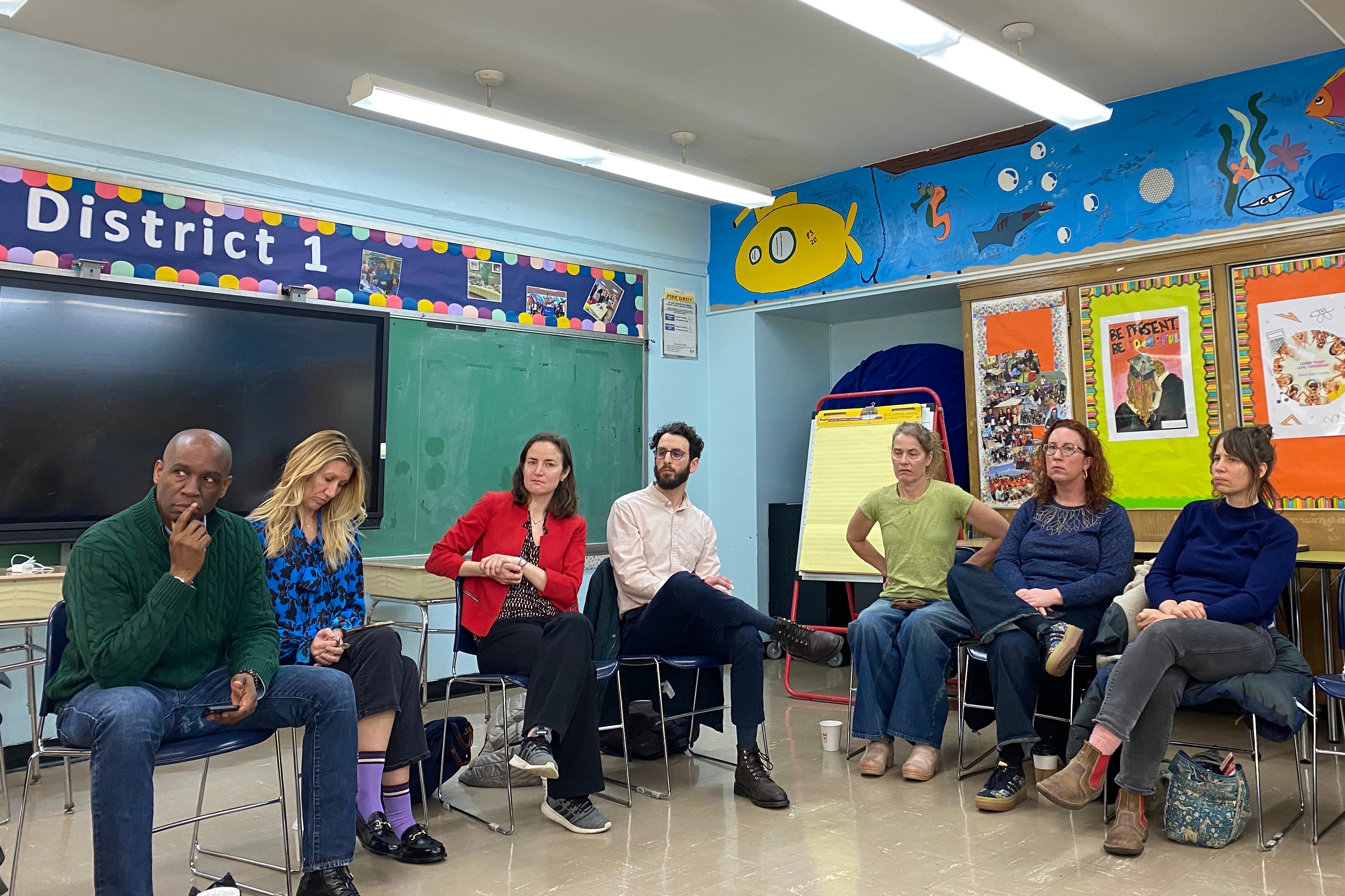 A room full of adults sitting in chairs in an L shape in a colorful classroom.