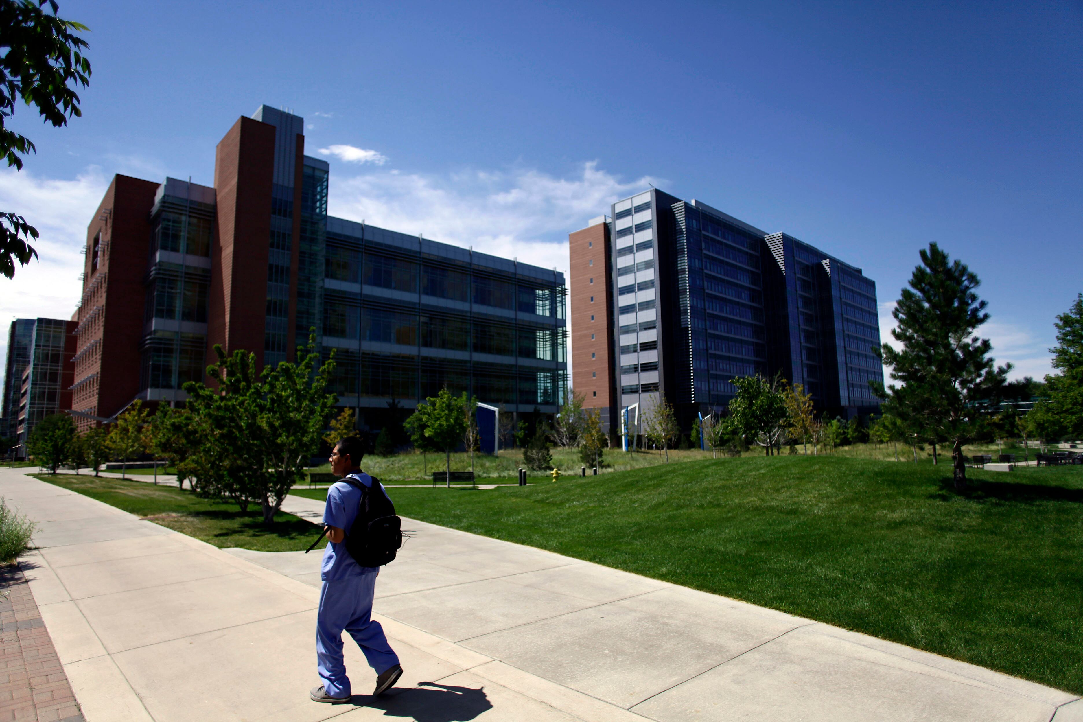 A person in scrubs and wearing a backpack walks down a sidewalk in front of a large medical building.