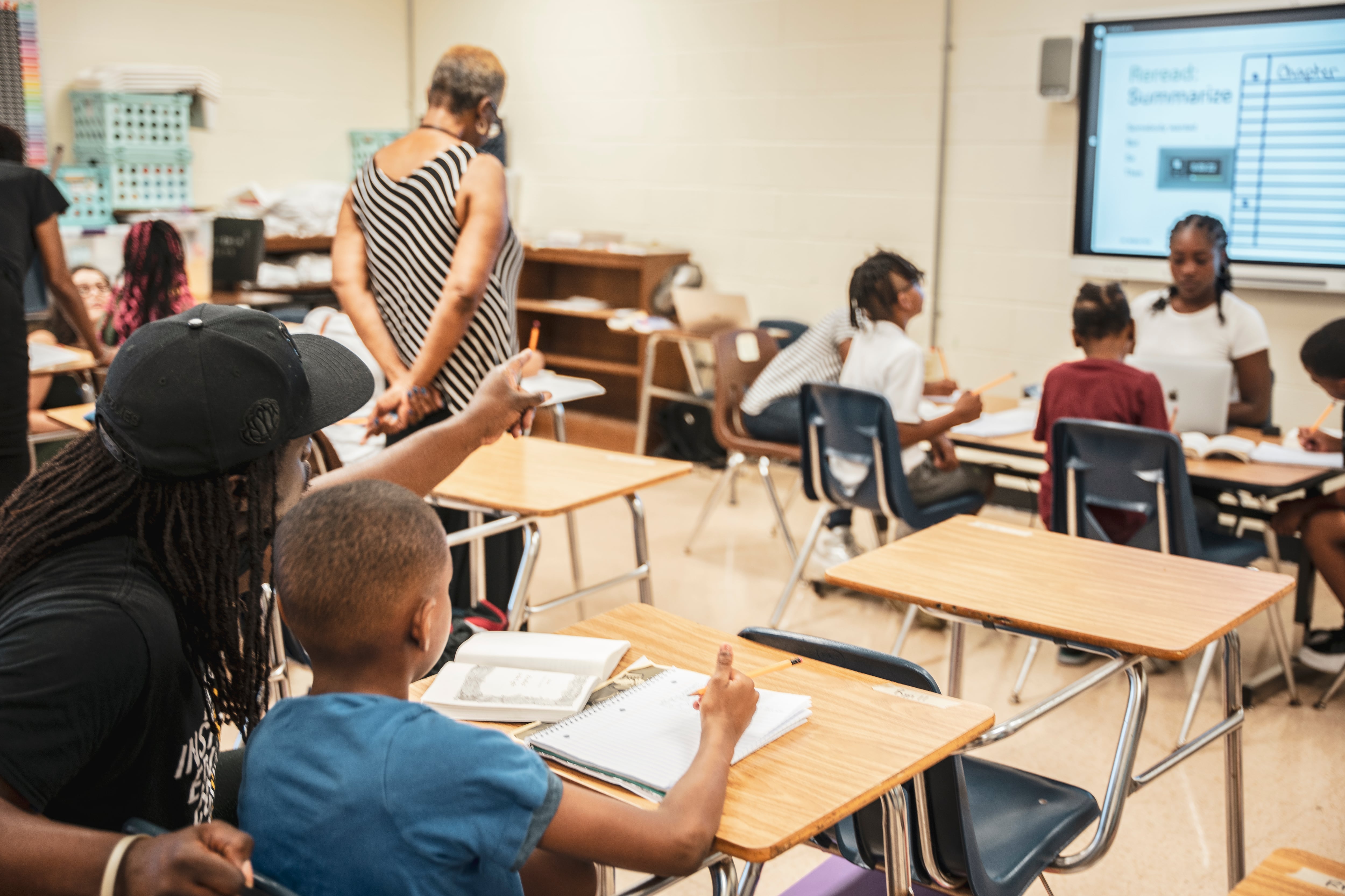 Three adults work with young students at their desks in a classroom with a projector screen on the back wall.