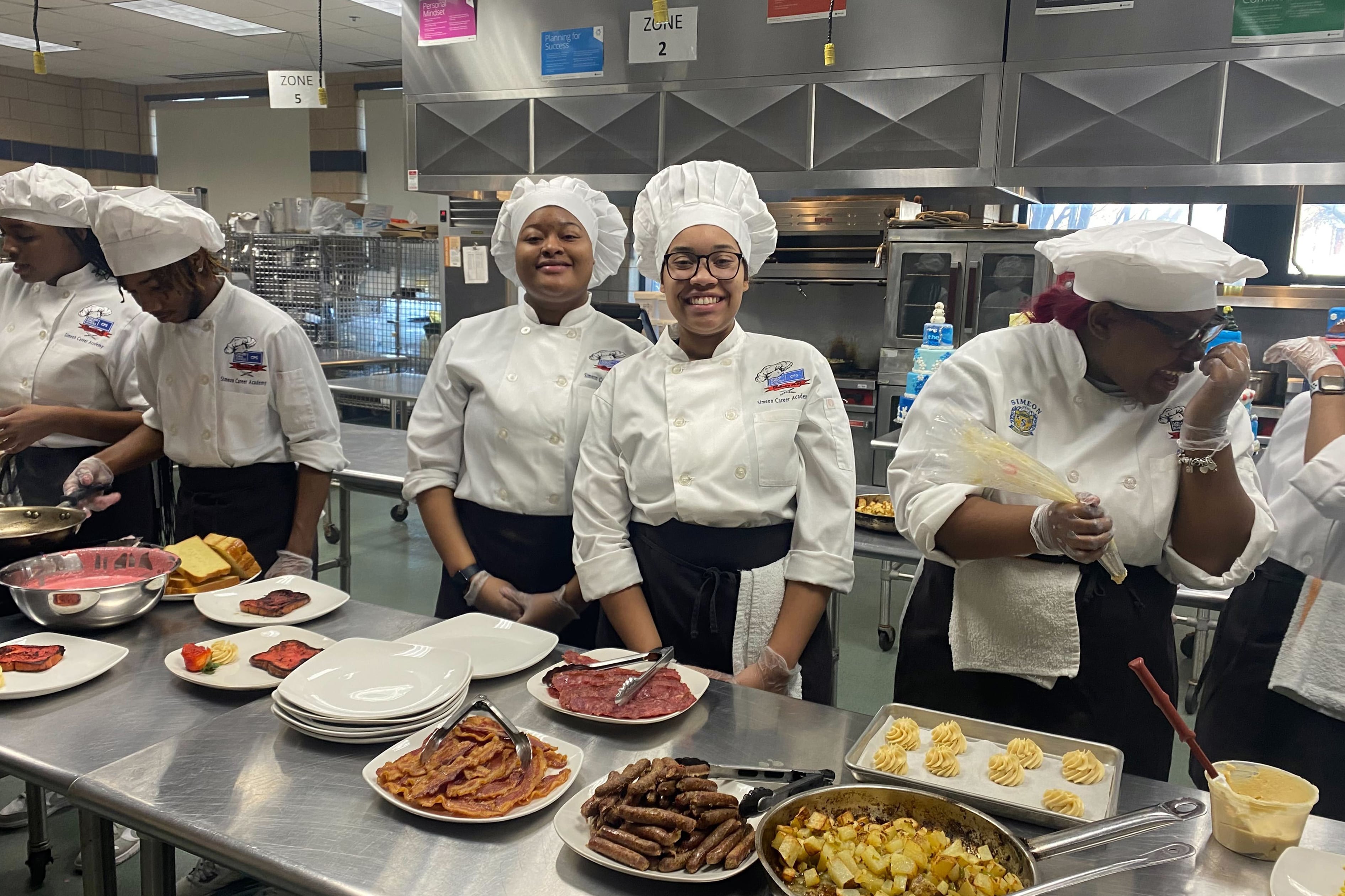 Six high school students wearing white chef coats and hats stand behind a metal table full of food.
