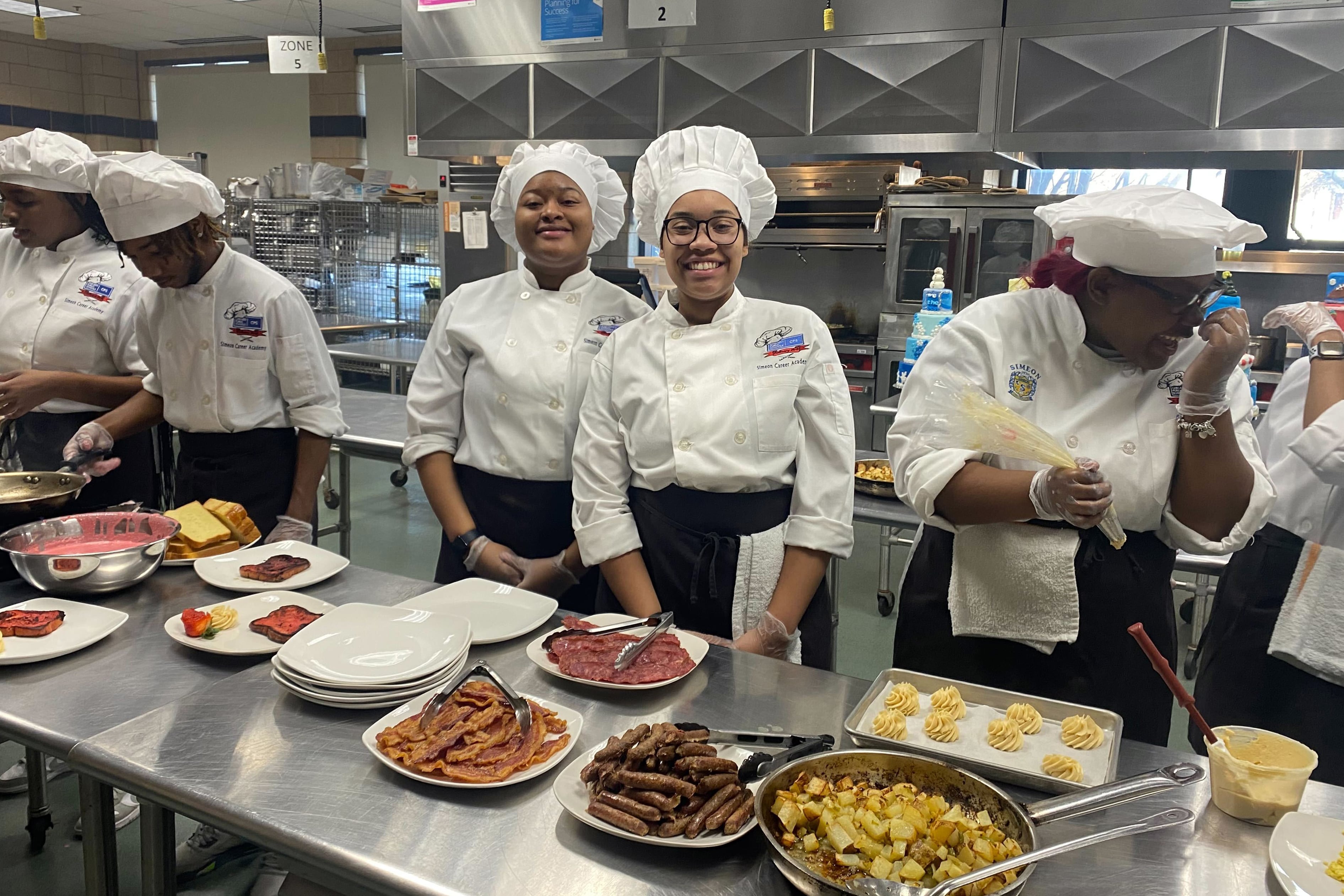 Six high school students wearing white chef coats and hats stand behind a metal table full of food.