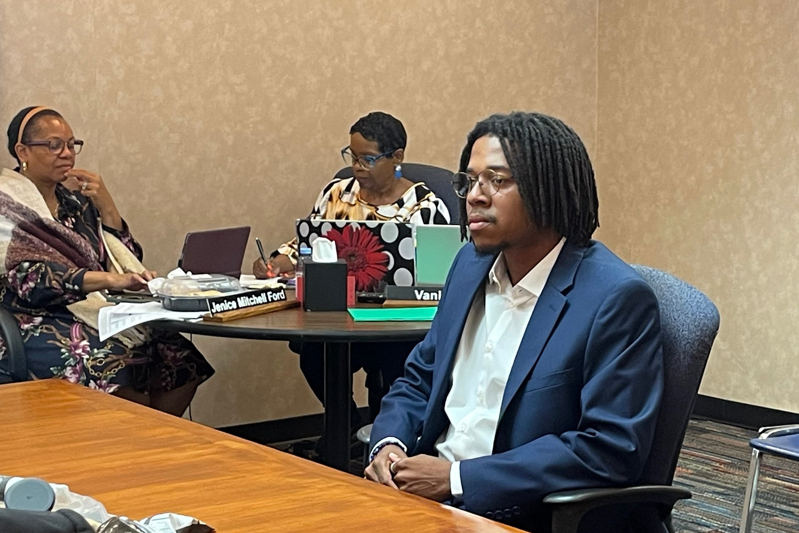 A Black man in a suit sitting in a chair at a wooden table with two people sitting in the background in a conference room.