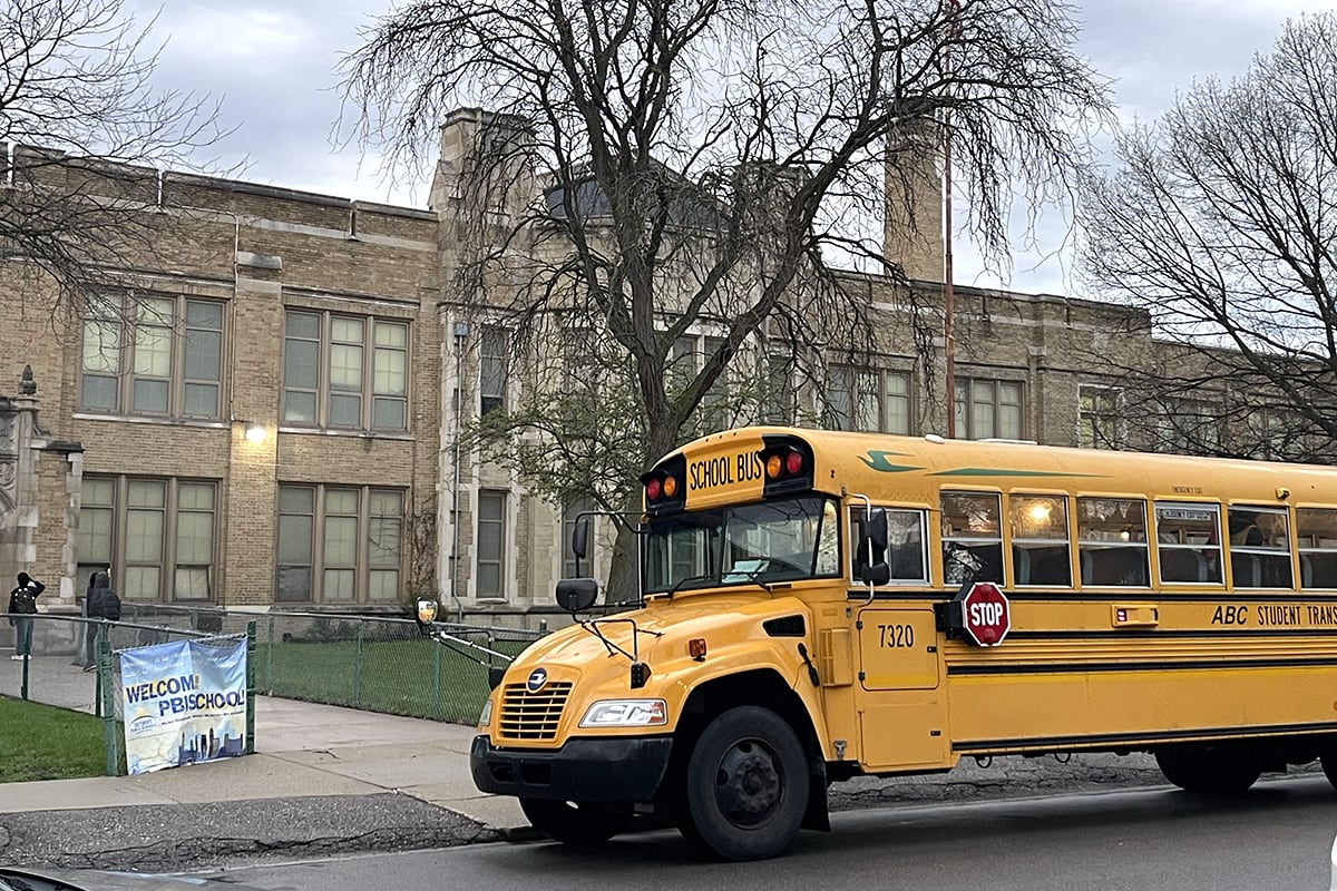 A photograph of a yellow school bus parked in front of a large school building on a cloudy day.