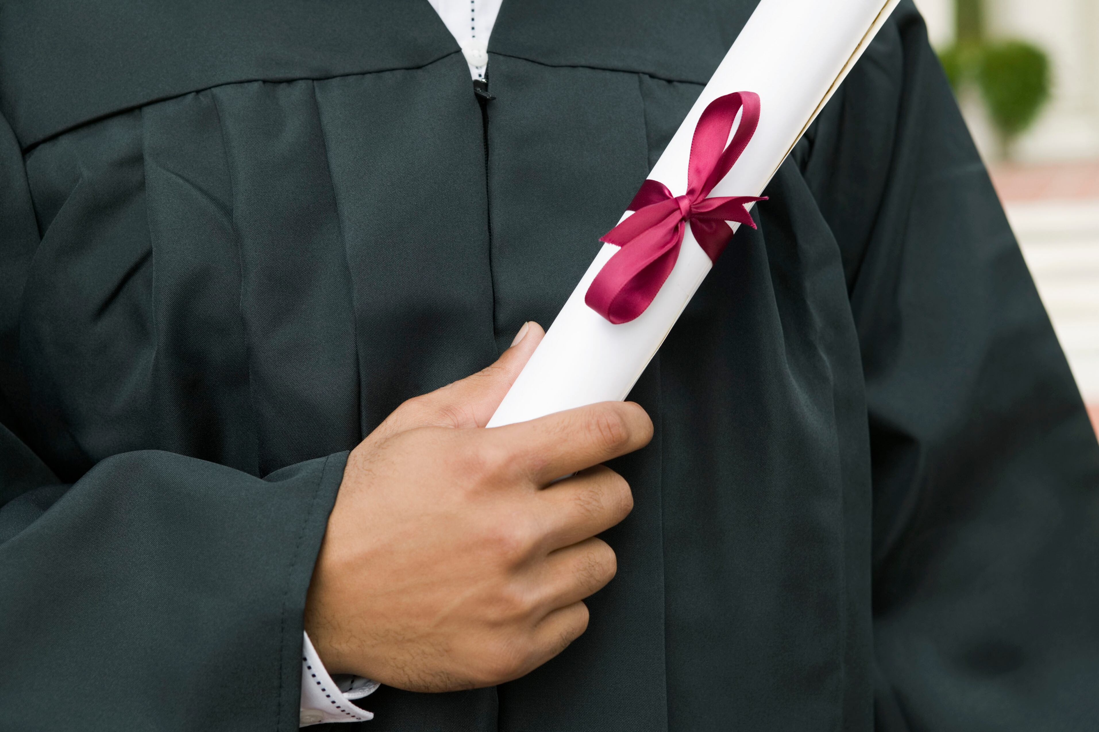 A close up of a hand of a high school graduate holding a white diploma with a red ribbon against their gown.