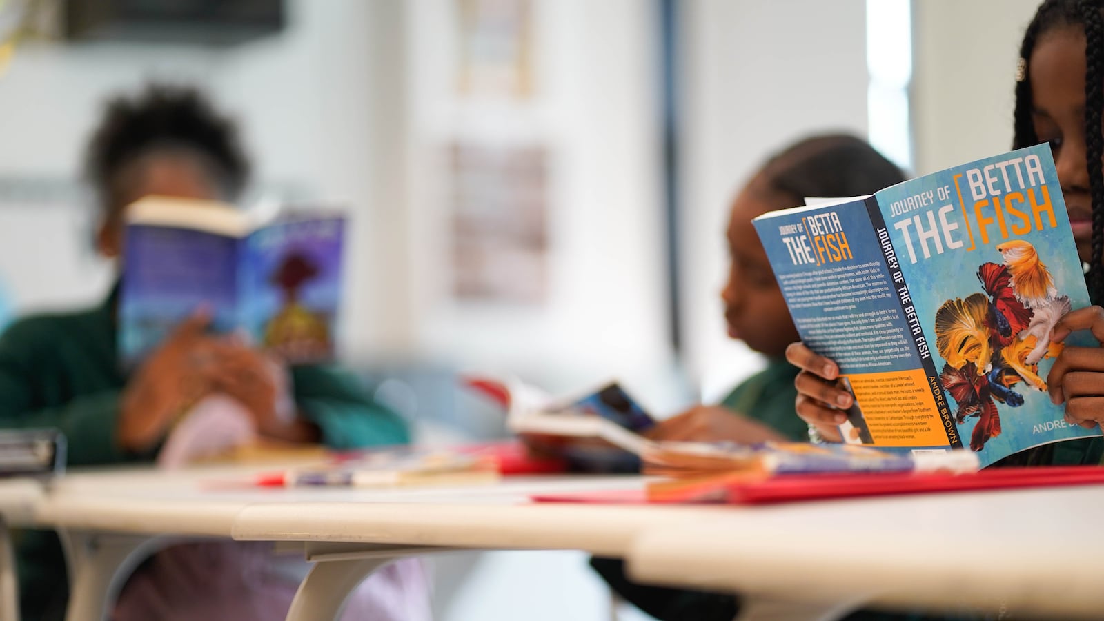 Three students sit at desk in a classroom reading books.