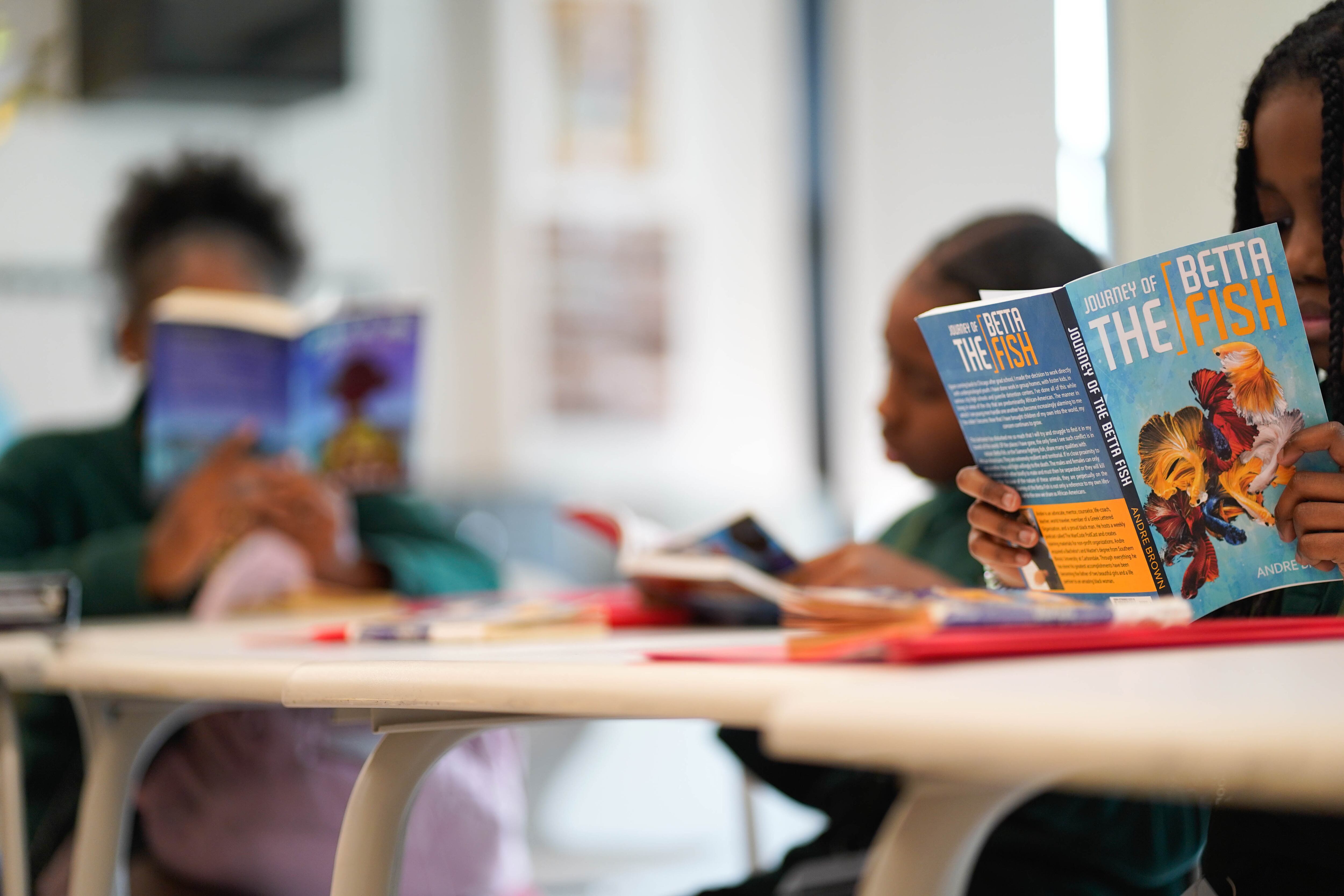 Three students sit at desk in a classroom reading books.