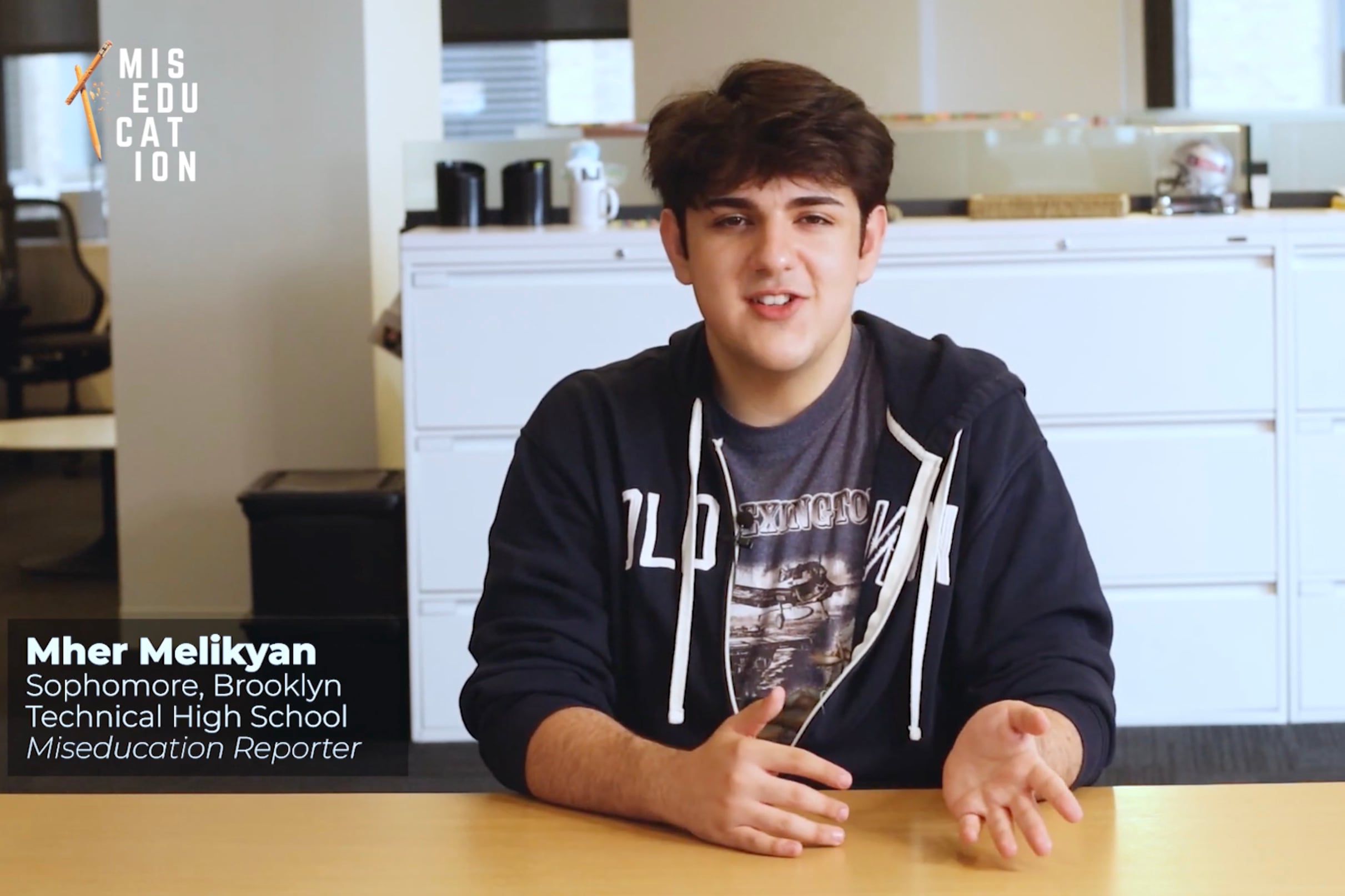 A screenshot of a high school student with short dark hair and wearing a hoodie sitting at a wooden desk with an office view in the background.