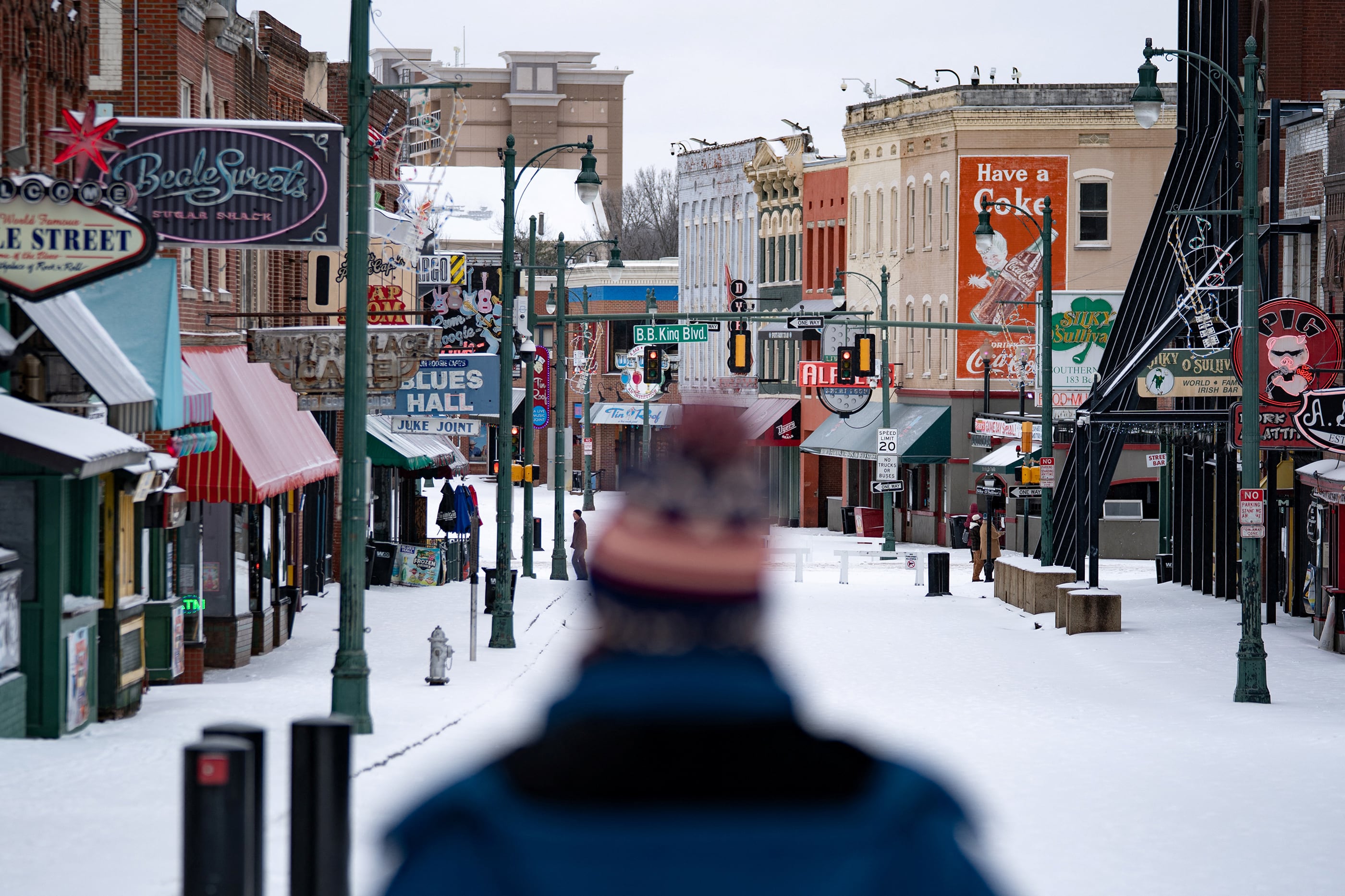 A photograph of a street filled with different shops and businesses covered in snow and the back of a person's head is right in the middle of the frame.