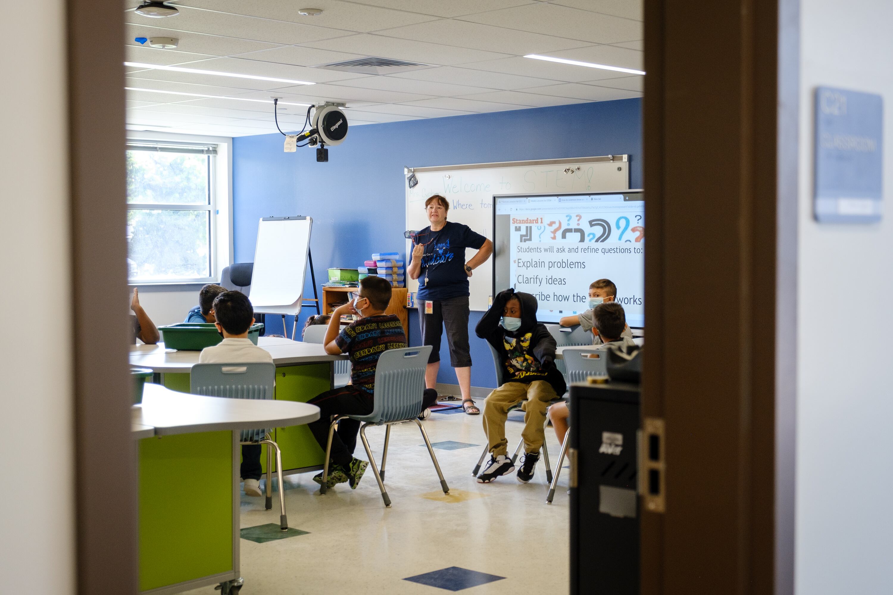 A teacher speaks to her students, wearing masks, during a STEM class. The classroom is partially obscured by the doorframe.