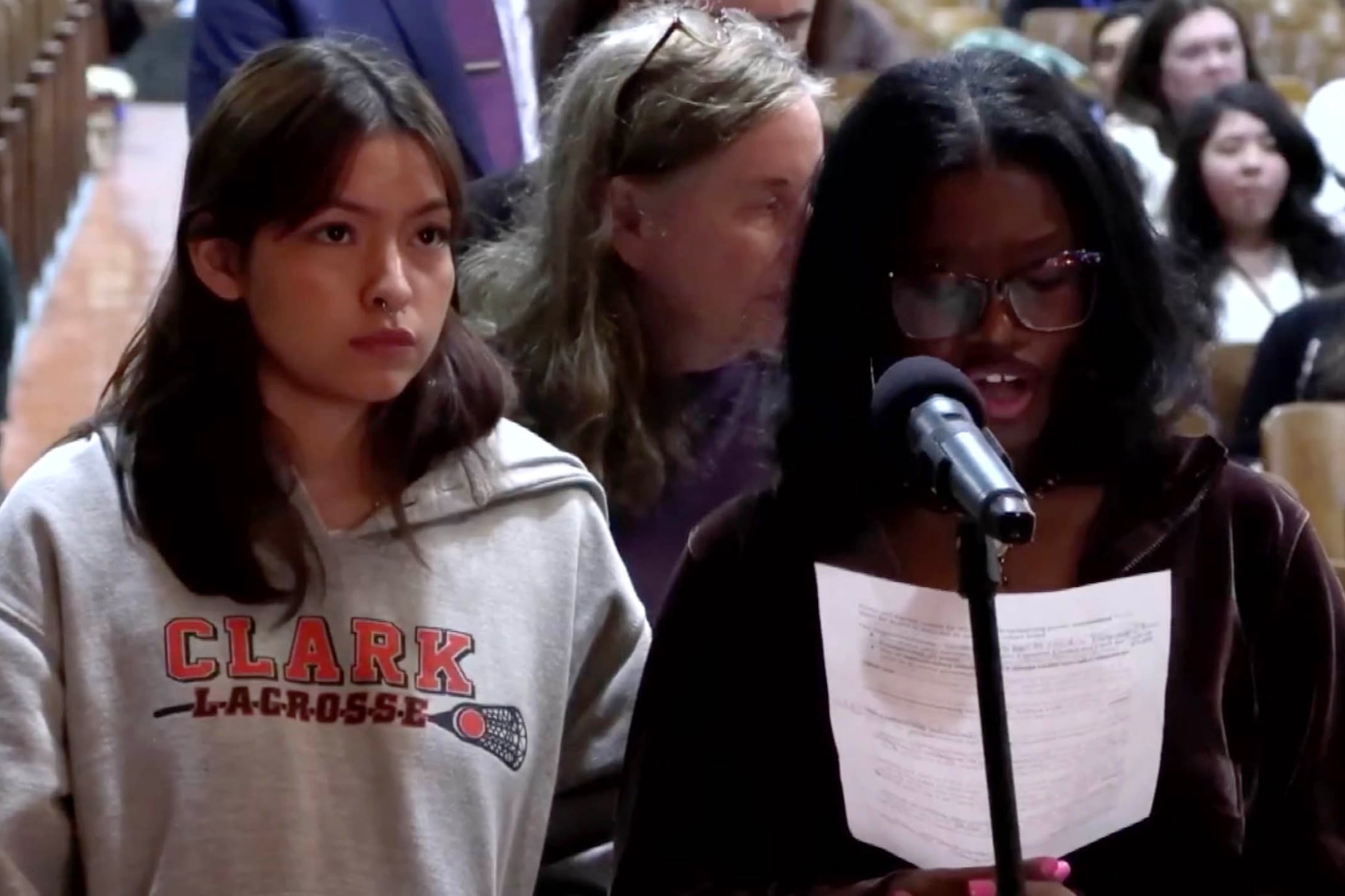 Two students with medium length hair stand next to each other while the student on the right reads from a piece of paper into a microphone with people sitting behind them.