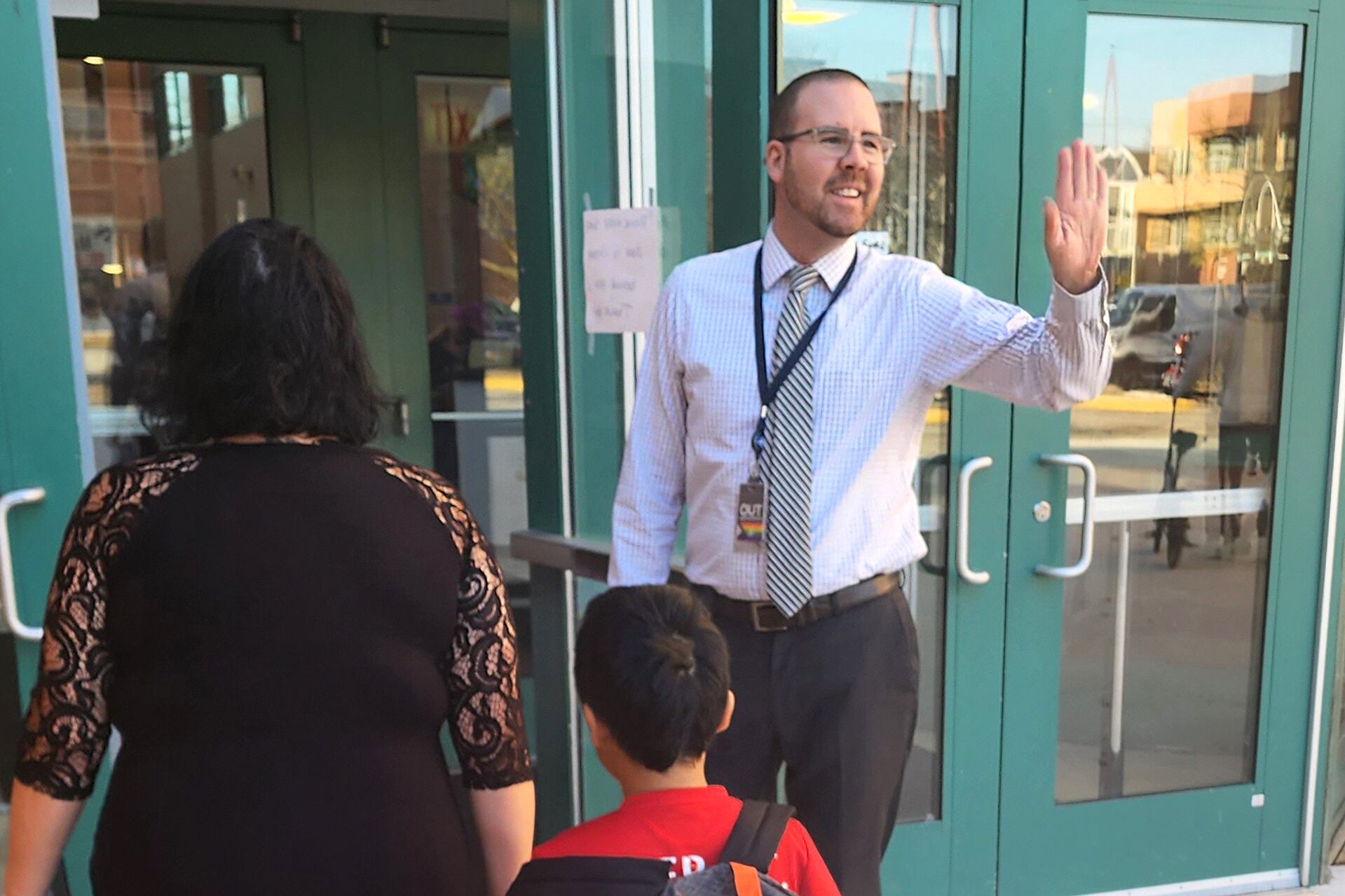 A man wearing a white shirt stands next to the front entrance to a school building while an adult wearing a black dress and a young student wearing a red shirt and a black and orange backpack walk by.