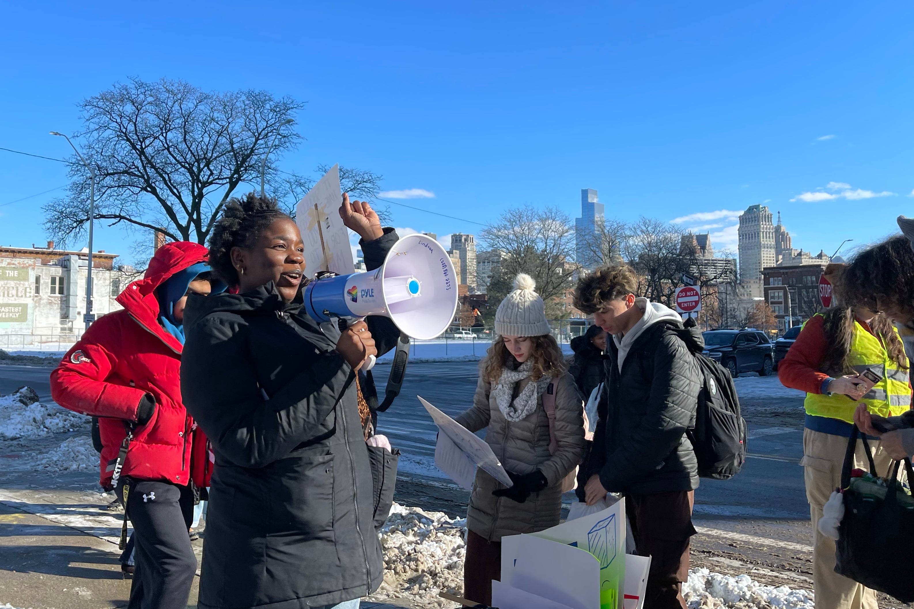 A photograph of students holding signs in the background while one Black student speaks from a microphone outside on a cold winter day.
