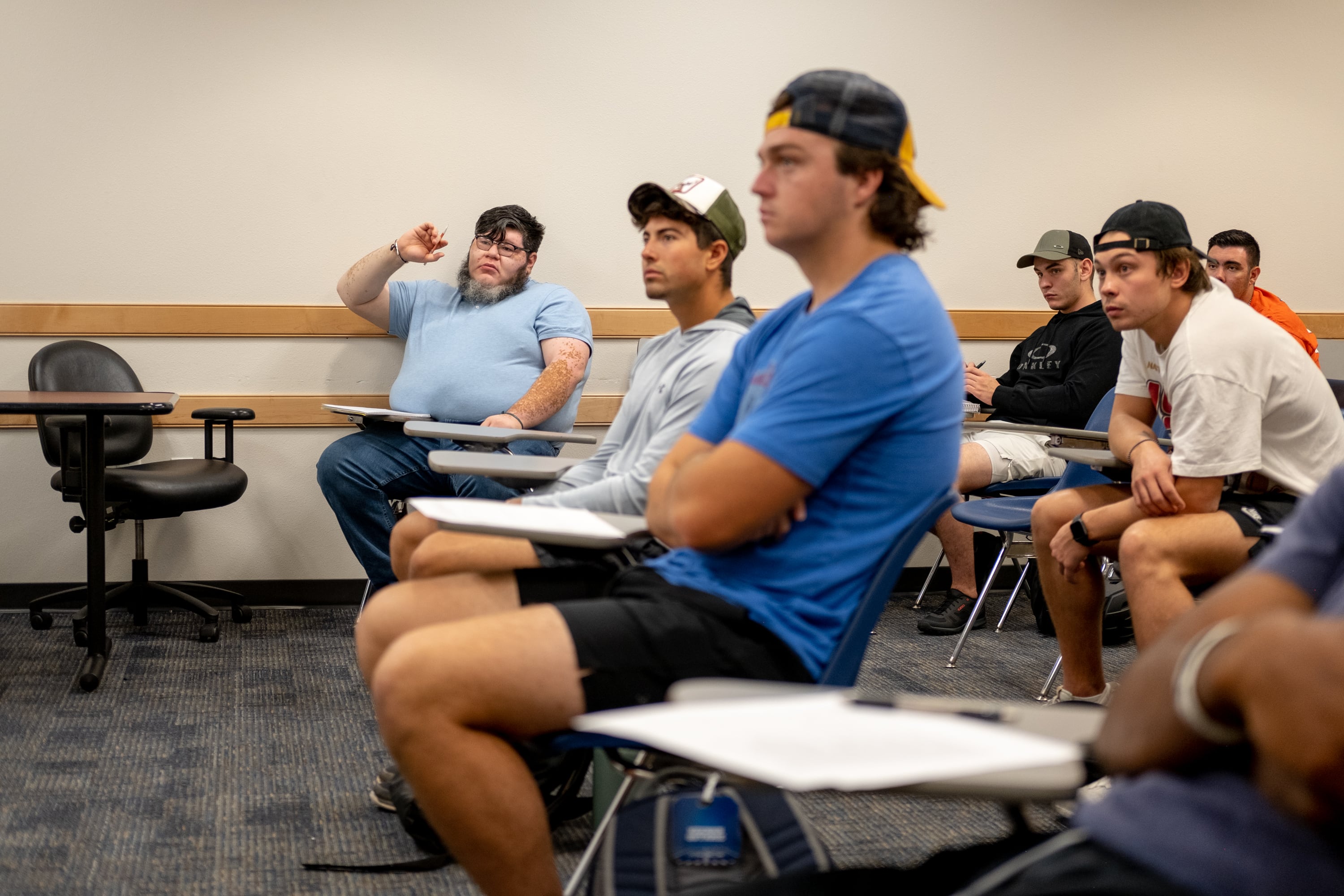 A student raises his hand in the classroom amongst other students sitting at their desks.